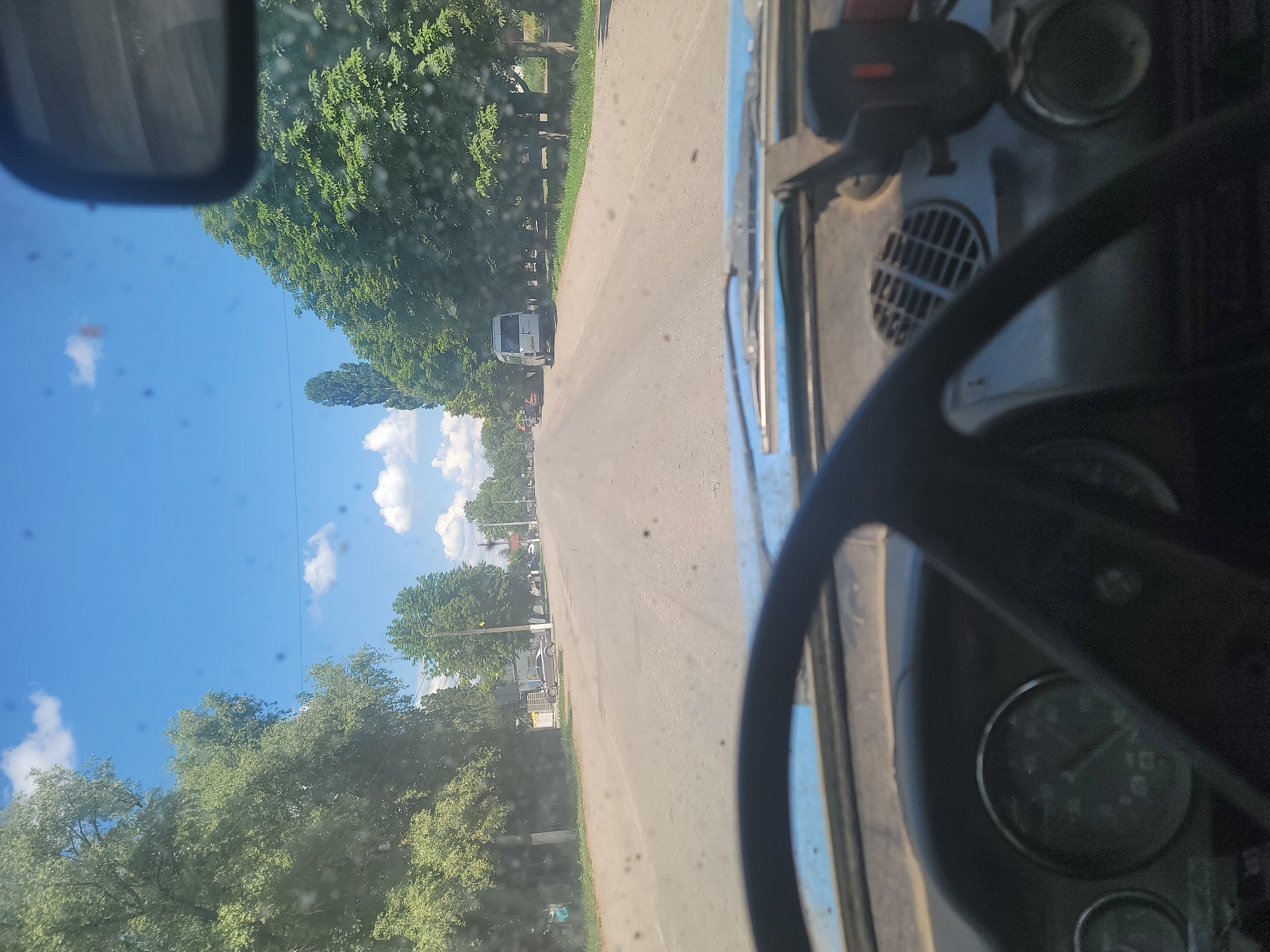 The image is taken from inside a vehicle, looking forward through the windshield. The perspective is from the driver's seat, with the steering wheel and part of the dashboard visible in the foreground. The road ahead appears to be asphalt and stretches into the distance. On either side of the road, there are trees with green foliage, suggesting a rural or suburban setting. A few buildings and parked vehicles, including a white minivan, can be seen along the roadside. The sky is blue with scattered white clouds. The scene is consistent with a drive through Pereshchepyne, Ukraine, given the context. The image is slightly dusty, with several small dark spots visible on the windshield.