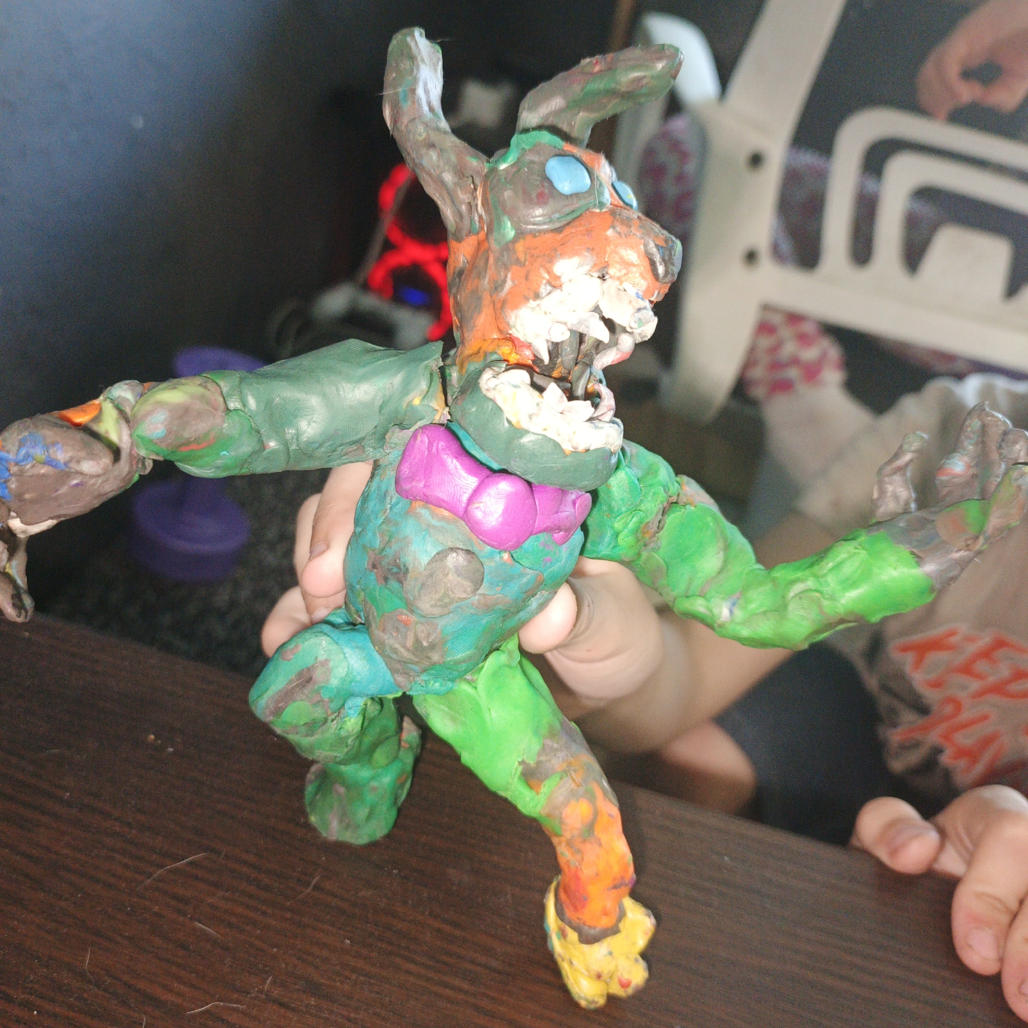 In the foreground, a child's hands are seen holding and manipulating a colorful clay figurine on a dark wooden surface. The figurine, an anthropomorphic creature with rabbit-like ears, is made of various colored clay, including green, orange, blue, pink, and yellow. It has large, bright blue eyes, a gaping mouth with visible teeth, and a pink bow tie or similar adornment on its chest. The creature appears to be in a dynamic pose, with its arms outstretched and legs bent.
In the background, out of focus, a person is partially visible, wearing a grey t-shirt with red text that appears to read "KEEP PLAY." The setting seems to be indoors, possibly a home, given the presence of a white plastic chair with patterned fabric underneath it. The lighting suggests it could be daytime, but the overall ambiance is somewhat dimly lit, with a hint of red light coming from an unseen source on the left. The image captures a moment of creative play and crafting, highlighting the interaction between the child and their handmade toy. The context provided of Vyshgorodok, Russia, adds a geographical detail to the scene.