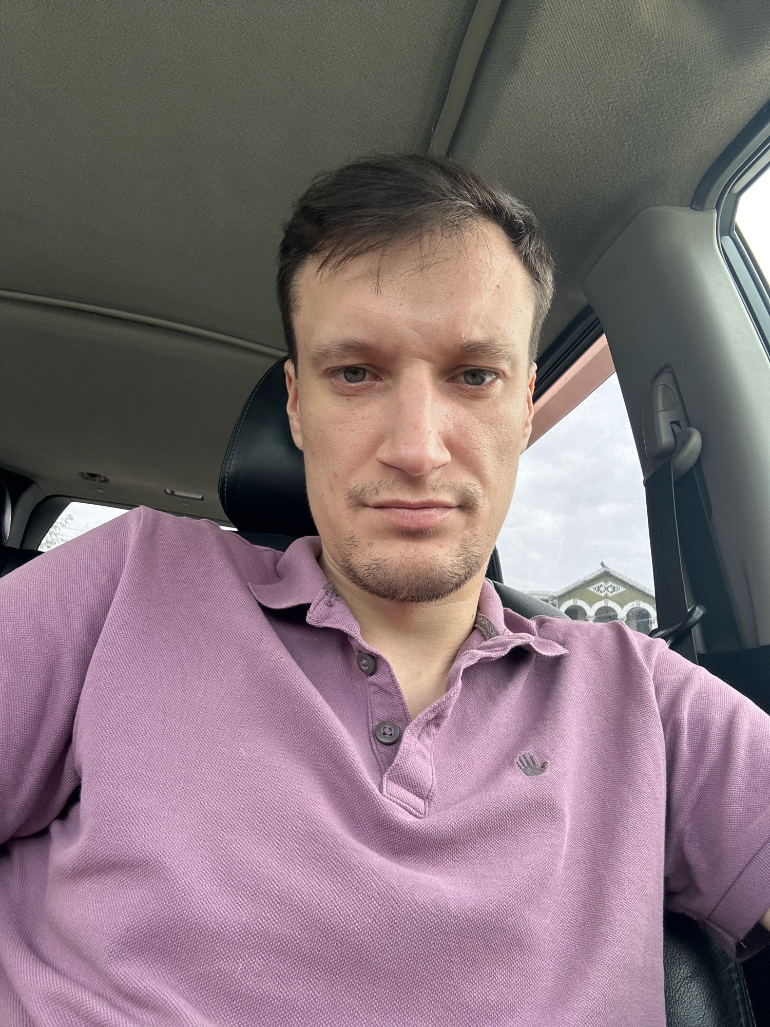 A man is taking a selfie inside a car. He is wearing a purple polo shirt. The background is slightly blurred but shows a building with arched windows and cloudy skies. The car interior is visible, including the headrest and ceiling.
