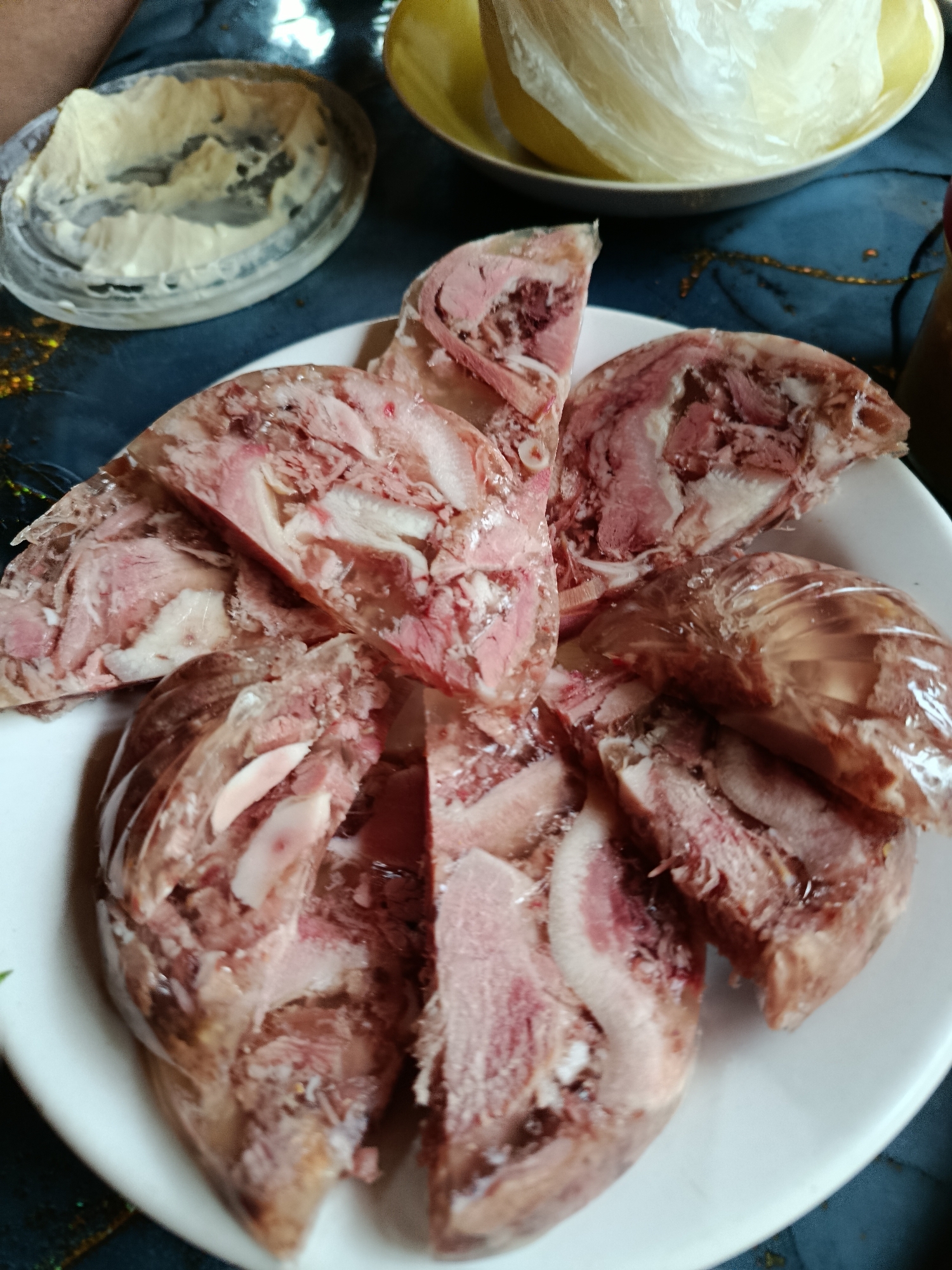 A plate of sliced jellied meat, likely brawn or kholodets, is presented on a white plate. The meat is a mix of pinkish-red muscle and white fat, suspended in a translucent, gelatinous substance. Some pieces show what appear to be bone fragments. The plate is situated on a dark blue tablecloth, which has some shimmering gold flecks. To the left of the plate, a small, shallow glass dish contains a pale yellow, creamy substance, possibly butter or a sauce. In the background, a yellow bowl is partially visible, containing an item wrapped in clear plastic. The overall scene suggests a home-cooked meal setting, possibly in Верея, Russia, given the context provided.