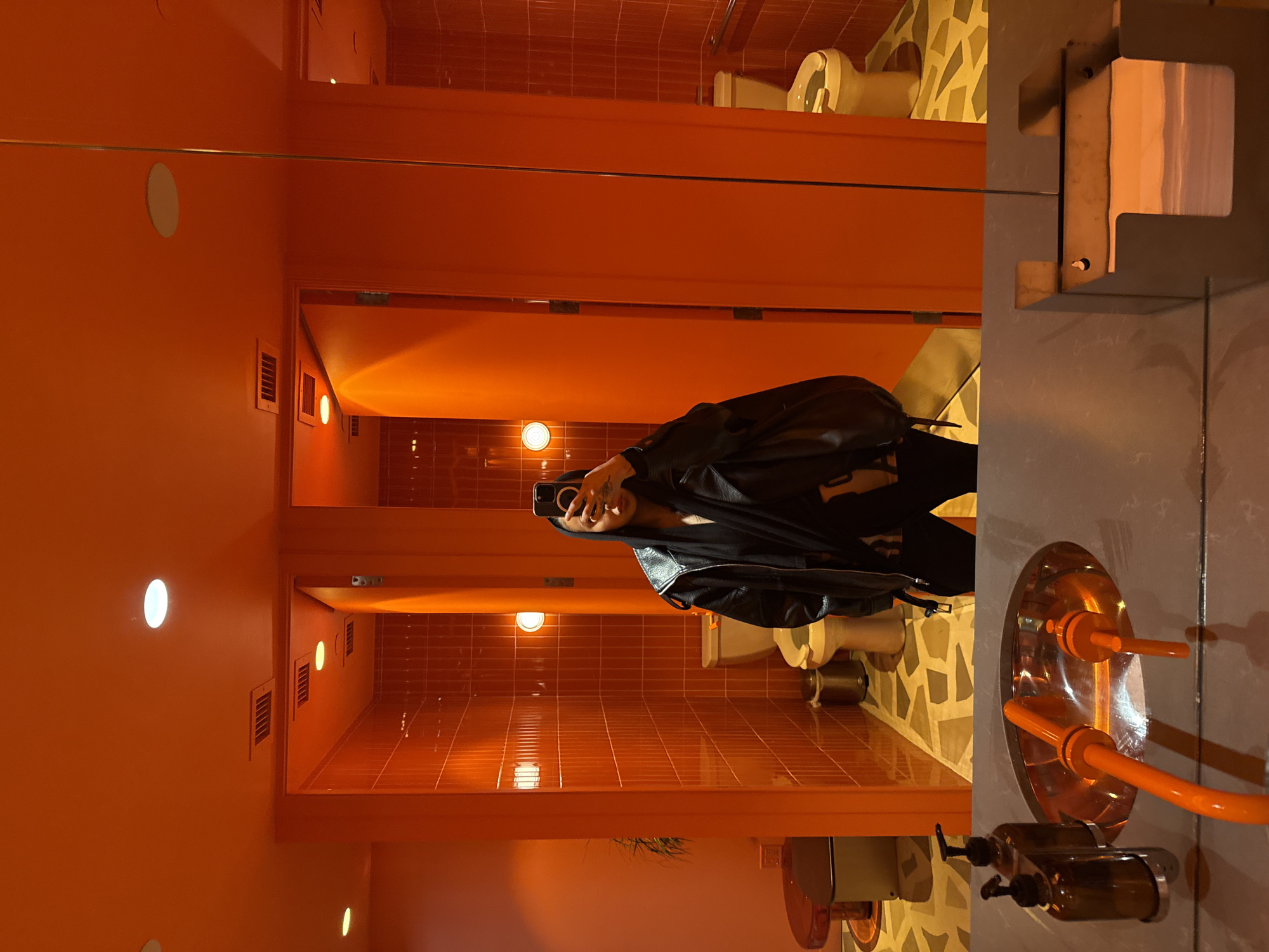 A person in a black leather jacket is taking a selfie in a public restroom. The restroom has an orange color scheme with a beige and white patterned floor. The restroom has multiple toilet stalls. In the foreground, there is a sink with vintage-style plumbing fixtures. The person is holding a phone to take the picture in the mirror. The location is likely in a public space in Vancouver, Canada.