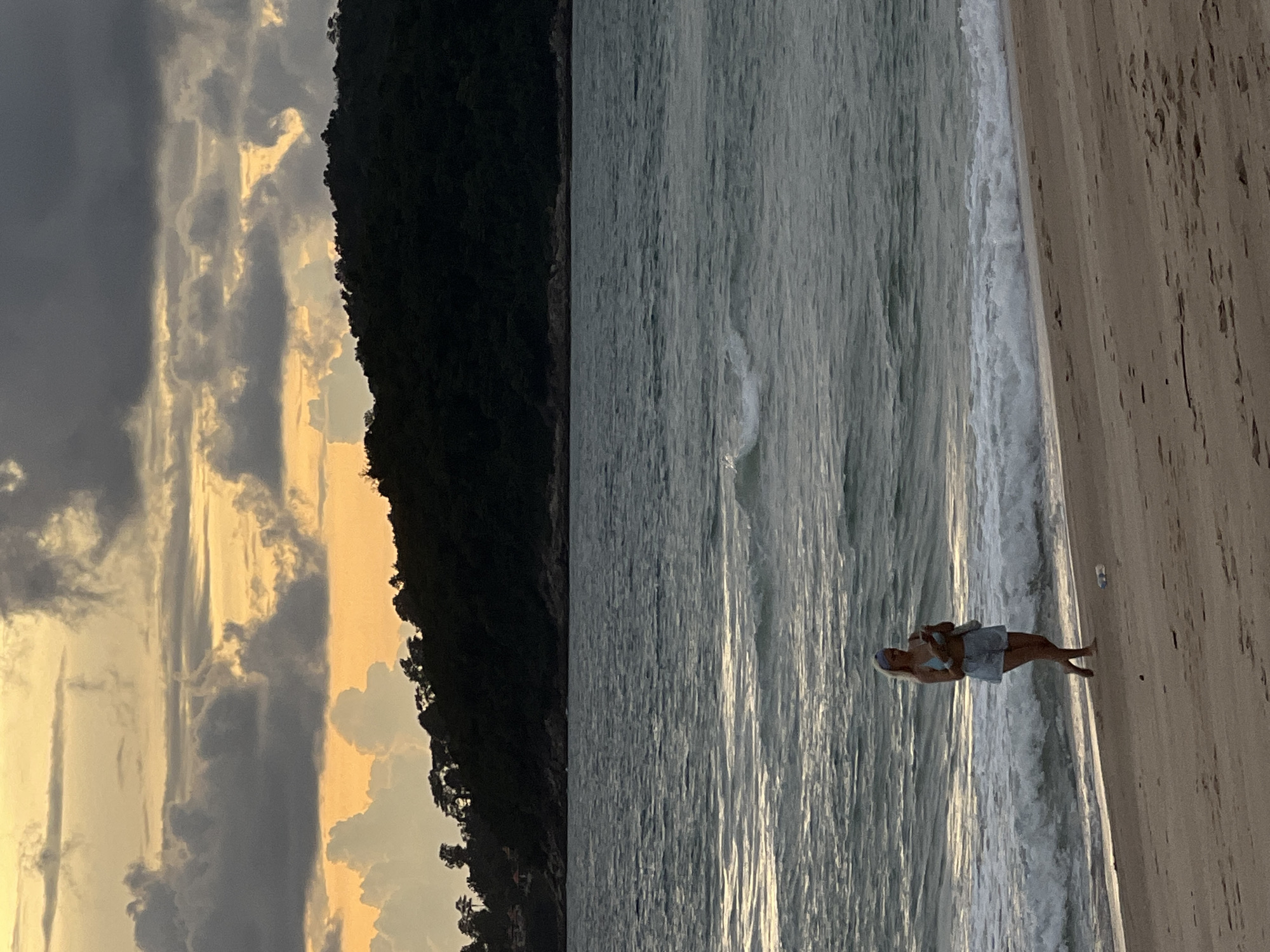 A woman is walking on a beach near the ocean at sunset. She is wearing a blue bikini top and denim shorts, and a blue headband. She is holding her phone in her right hand and looking down at it. The waves are gently crashing on the shore, and the sky is filled with dramatic clouds in shades of orange, yellow, and gray. In the distance, a dark, tree-covered hill rises above the horizon. The beach is sandy and mostly empty, with a discarded plastic bottle visible on the sand to the right of the woman.