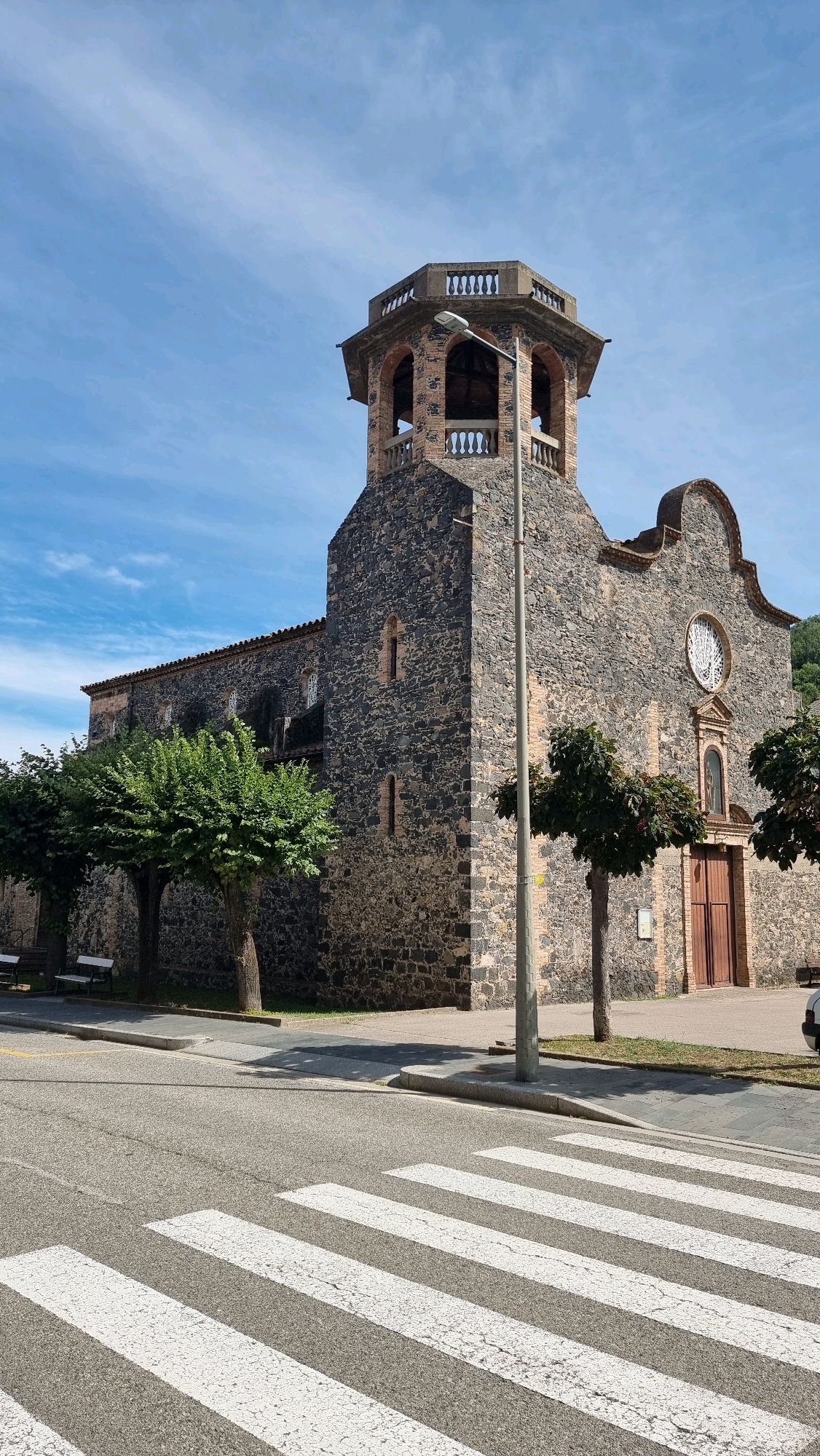 The image shows a stone church in Castellfollit de la Roca, Spain, on a sunny day with a blue sky and scattered clouds. The church has a distinctive bell tower with arches and a decorative railing. In front of the church, there is a street with a pedestrian crossing marked by white stripes. Several trees are visible along the sidewalk. The architecture appears old, made of dark stone with some lighter stone accents. A lamppost stands near the church entrance. The overall scene depicts a quiet, urban environment.