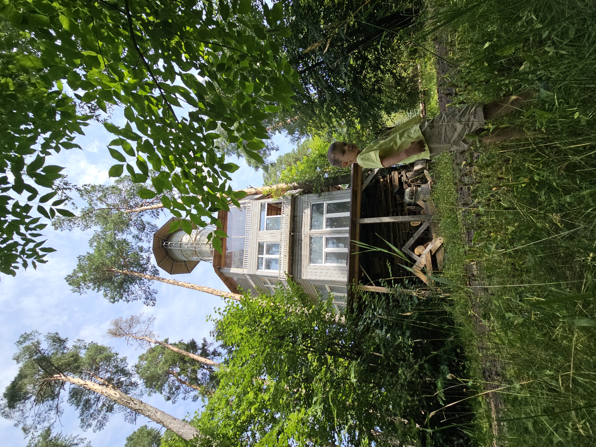 In the city of Ulitkino, Russia, a low-angle shot captures a unique wooden structure resembling a multi-story tower, possibly a treehouse or observation deck, nestled within a dense forest. The structure, clad in gray wood siding, features multiple levels with numerous windows, topped by a octagonal section with a metallic roof. Surrounding the tower are tall, slender pine trees reaching towards a bright blue sky dotted with white clouds, and lush green foliage from deciduous trees. In the foreground, a young boy, wearing a yellow t-shirt and khaki shorts, stands partially obscured by tall grass, looking towards the camera. The ground beneath the structure is visible, showing stacked firewood. The overall scene depicts a natural, somewhat rustic setting with a distinctive architectural element.