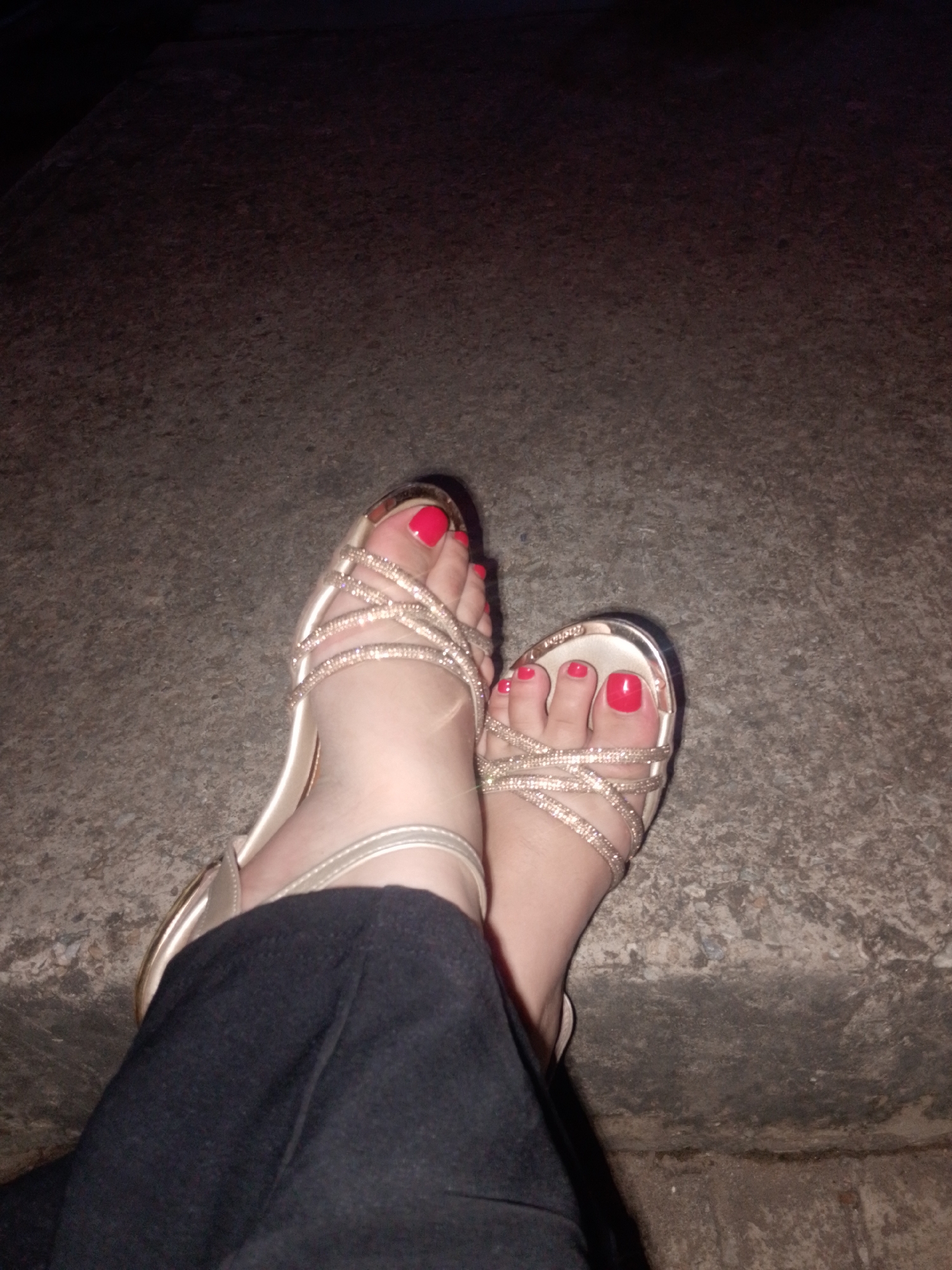 A low-light, high-angle close-up shot captures a person's feet, clad in fashionable footwear, resting on a textured, dark ground surface. The scene is dimly lit, suggesting it is nighttime or takes place in a poorly lit indoor or outdoor area in Juan Griego, Venezuela.
The primary subjects are the two feet, positioned casually with one slightly overlapping the other. Both feet are adorned with open-toed, high-heeled sandals featuring multiple crisscrossing straps embellished with what appears to be sparkling rhinestones or glitter. The toenails are painted a vibrant, glossy red or coral color. Part of a pair of dark-colored trousers, likely black, is visible around the ankles and lower legs.
The ground beneath the feet is a rough, uneven, dark gray or brown surface, possibly concrete or asphalt, showing signs of wear and age. The lighting casts subtle shadows and highlights on the sandals and feet, emphasizing the sparkling details of the straps and the sheen of the nail polish. The background is largely indistinguishable due to the low light and shallow depth of field, fading into deep shadow. No text is visible in the image.