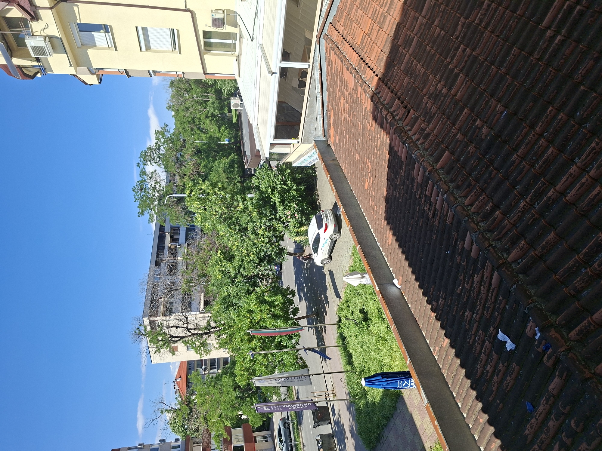 The image shows a view from an elevated position, likely a building in Plovdiv, Bulgaria. In the foreground, there is a red tiled roof. Below, a street is visible with a sidewalk lined with green vegetation. Along the sidewalk, several flags are mounted, including a Bulgarian flag. A white car is parked along the street. Trees line the street, partially obscuring buildings in the background. The sky is clear and blue.