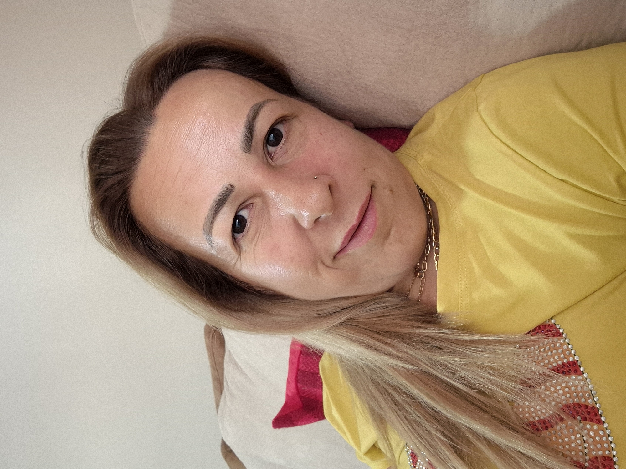 A medium shot, selfie-style photograph shows a woman with long, blonde hair, wearing a yellow t-shirt, and lying on her side on a couch. The woman is looking at the camera with a slight smile. Her hair is parted on the right side and falls over her left shoulder. She is wearing a gold chain necklace and has a small nose stud. The couch is covered in a neutral-colored fabric. A red pillow is visible behind her head. The background is a plain wall.