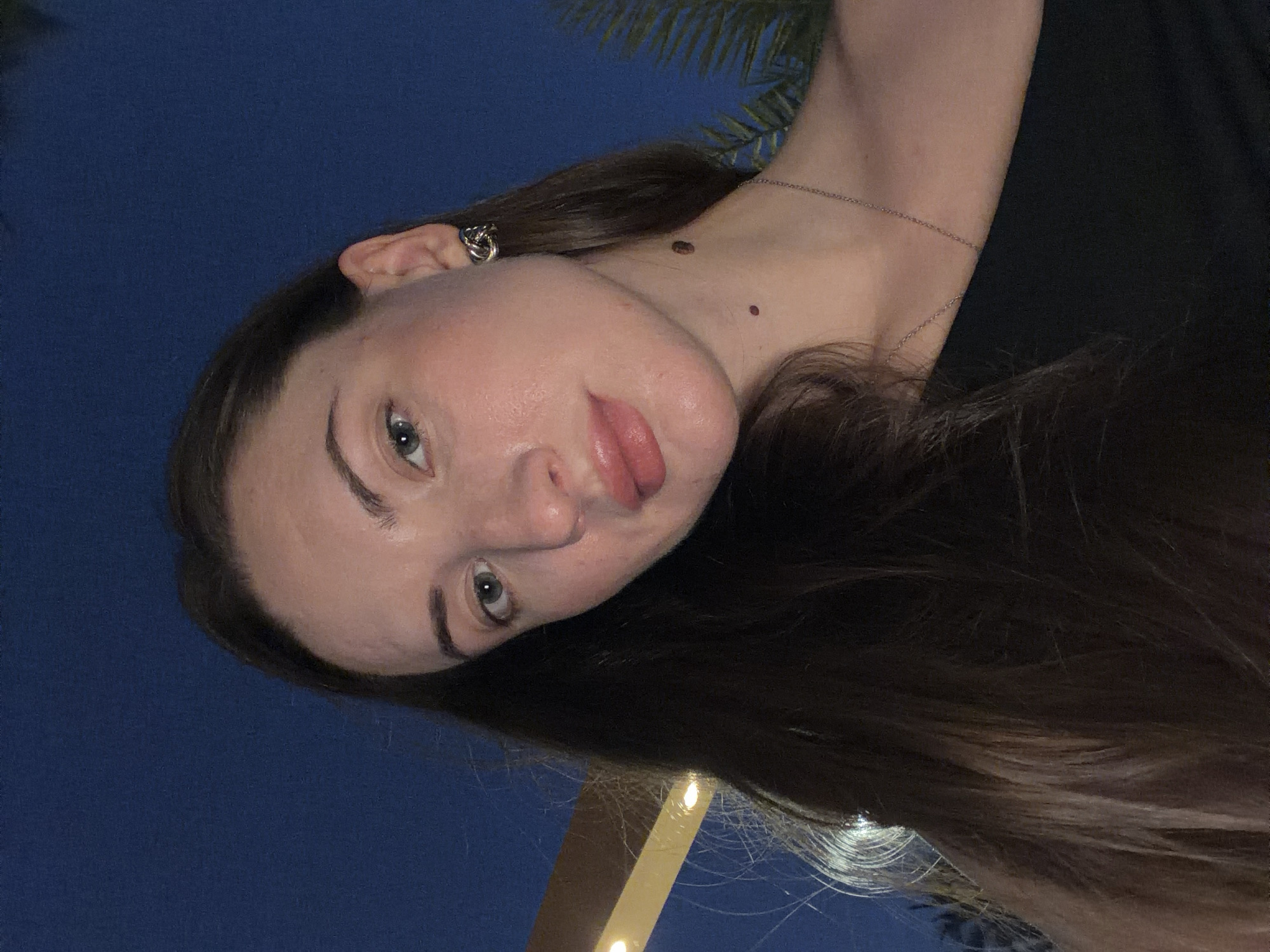 A close-up selfie captures a young woman with long, dark brown hair, a pale complexion, and light blue eyes. She is wearing a black off-the-shoulder top and a silver necklace. Her left ear is adorned with a knot-shaped earring. A small mole is visible on her neck below her jawline. The background is dark blue, suggesting it is nighttime. A blurry element of a palm tree and a lit structure with a yellow accent are visible on the left and right sides of the frame, respectively. The lighting suggests an outdoor setting in an urban or resort area at dusk or night.