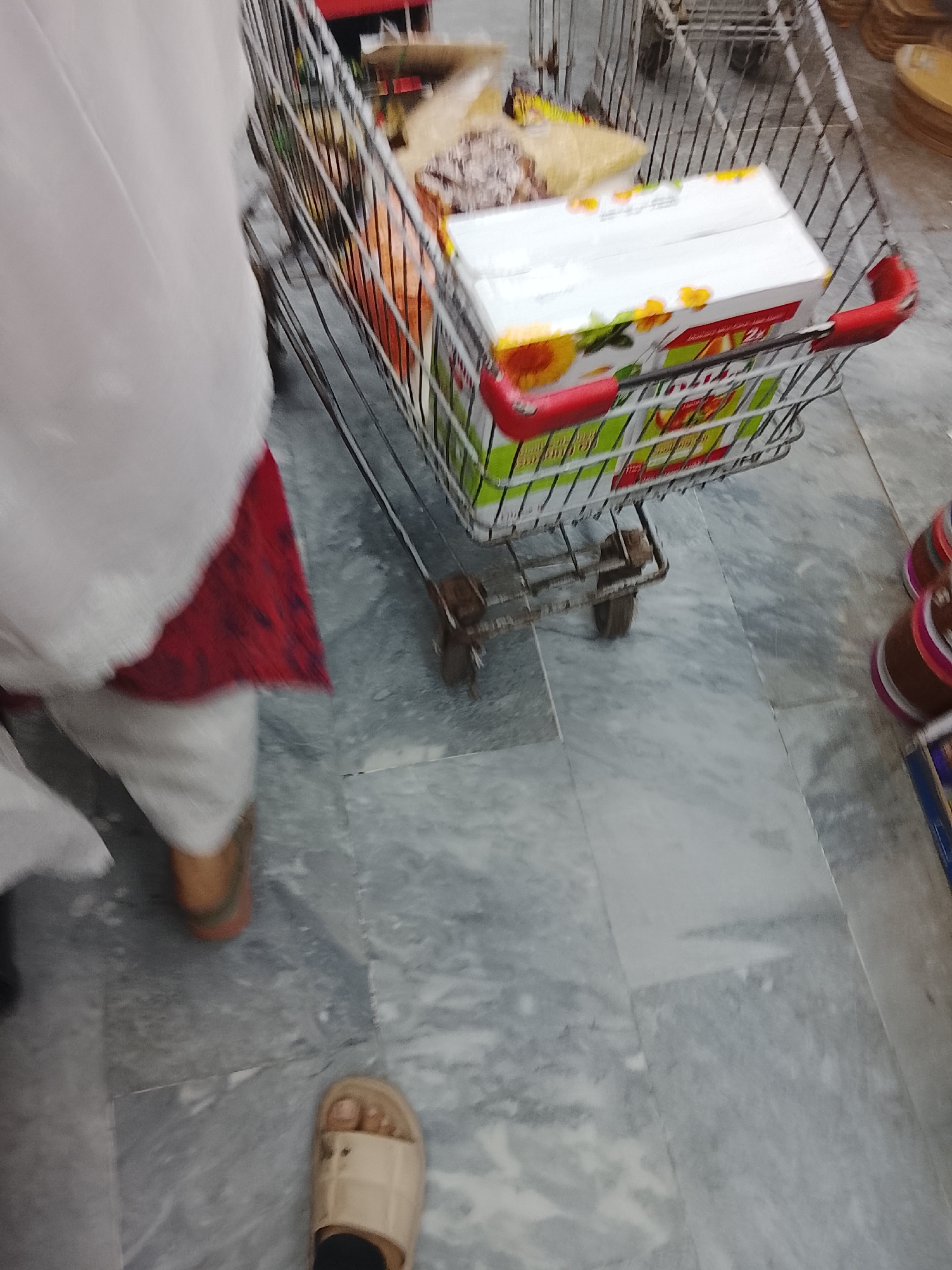 The image shows a person pushing a shopping cart in what appears to be a grocery store. The cart contains several items, including boxes of cooking oil and other packaged goods. The person is wearing a white headscarf, a red and white patterned garment, and off-white pants. Their feet are visible, wearing one tan sandal and one brown shoe. The floor is tiled with grey marble pattern tiles. The background shows other shopping carts and merchandise. Given the prompt, the location is likely a grocery store in Gulgasht Colony, Pakistan.