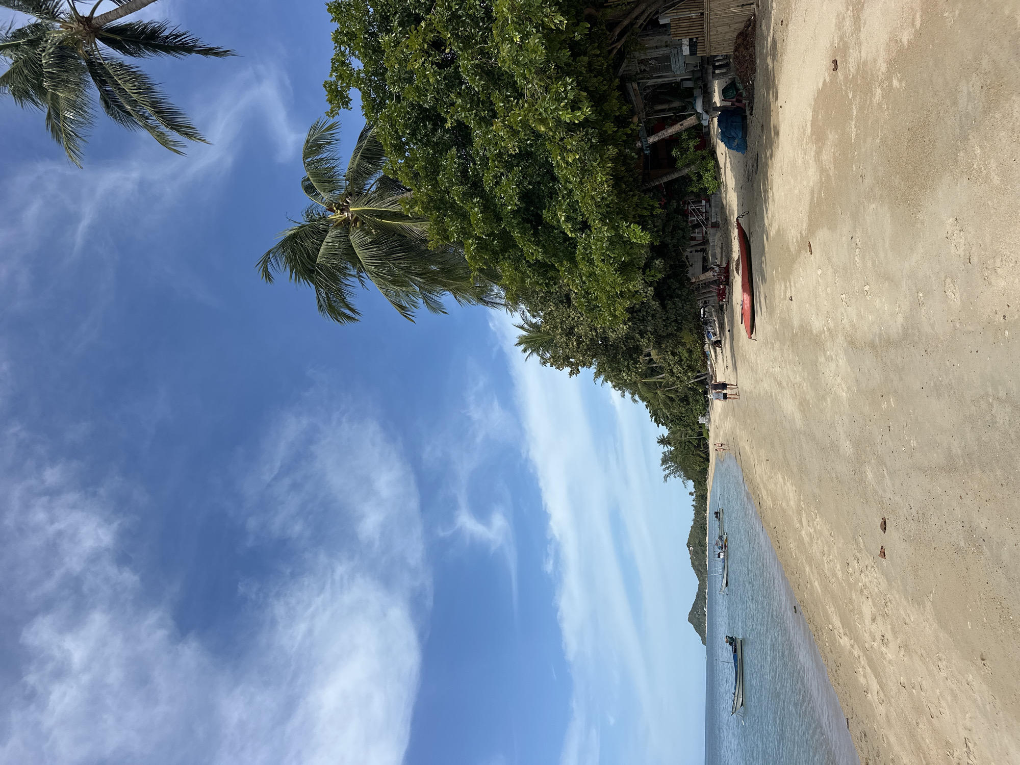 This image depicts a beach scene in the Ko Samui District of Thailand. The sand is light brown and textured, stretching from the foreground towards a line of lush green trees and palm trees. In the distance, two small, hilly islands are visible in the blue ocean, which is calm and reflects the bright sky. Several fishing boats are anchored in the shallow water. Two people are walking along the shore in the middle ground. Closer to the foreground, a red kayak rests on the sand. Behind the trees, there are glimpses of what appear to be simple structures. The sky is a vibrant blue with scattered wispy white clouds.