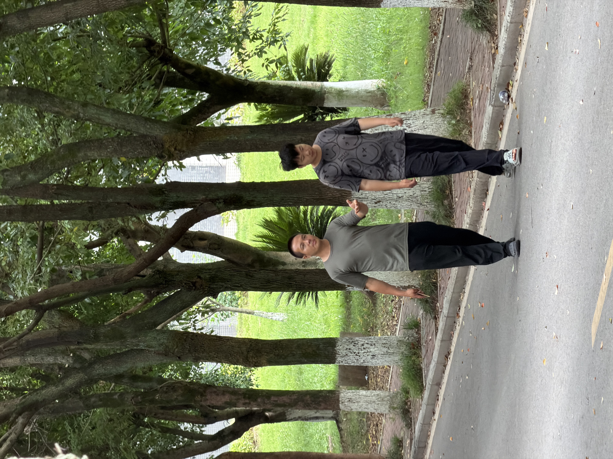 In the city of Jiangjin, China, two men are walking on a paved road bordered by a brick pathway and a grassy area with trees. The man on the left is wearing a dark green t-shirt and black pants, and he is holding a thin, light-colored object in his right hand. The man on the right is wearing a dark grey t-shirt with a repeating smiley face pattern and black pants. He is looking downwards. The trees have white paint around their bases. The environment is outdoors with green grass and foliage.