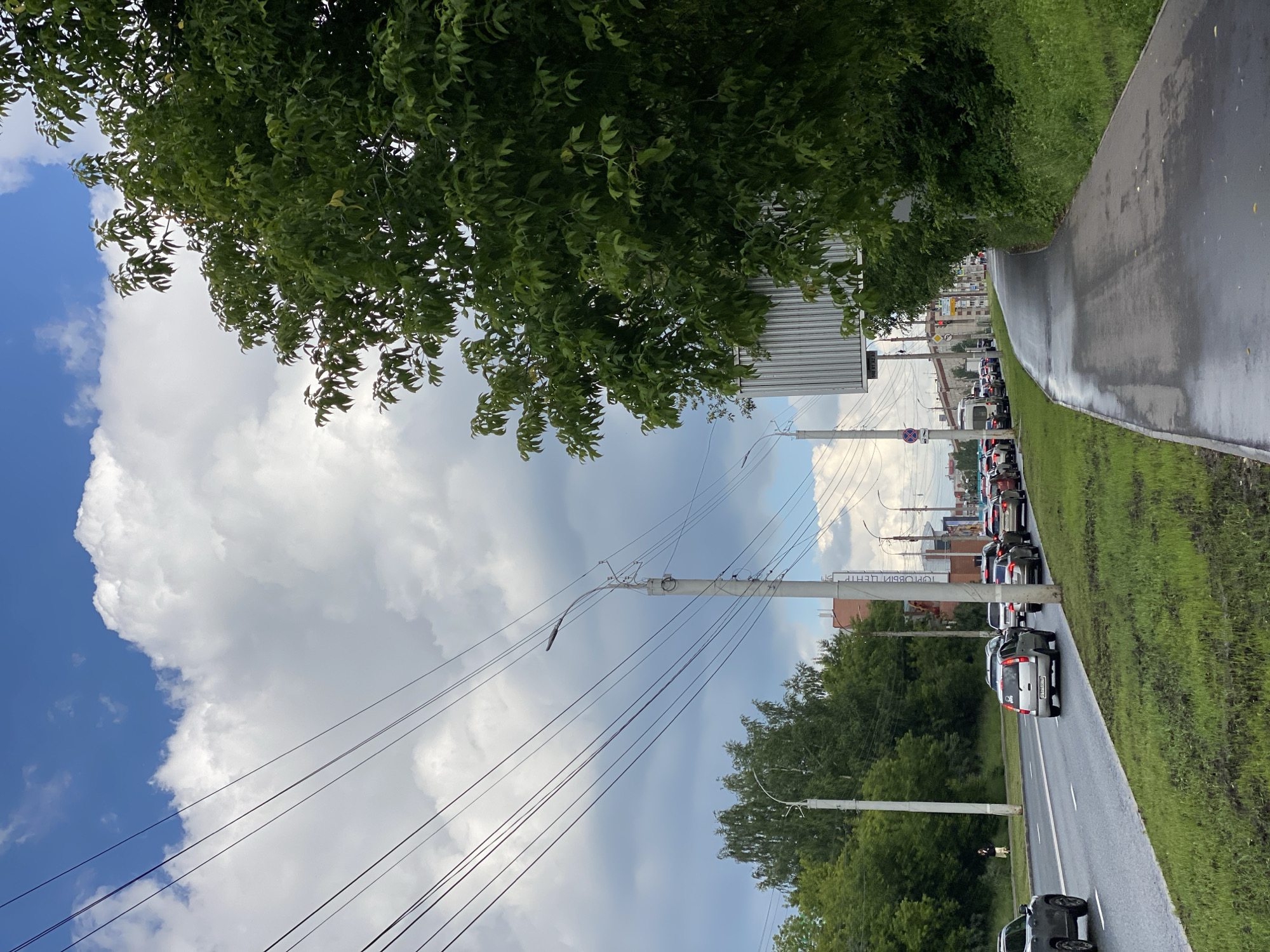 The image depicts a roadside scene in a city. A wet asphalt sidewalk runs along the right side of the frame, bordered by lush green grass and trees. Beyond the grass is a multi-lane road with several cars in traffic, indicating a moderate amount of congestion. Overhead, power lines crisscross the sky, which is a mix of blue and white clouds, suggesting possibly overcast or partly cloudy weather. Streetlights and utility poles are visible along the roadside. In the distance, buildings and trees line the street. One person is visible walking on the sidewalk on the left side of the road. The overall impression is an urban environment on a day with dynamic weather.