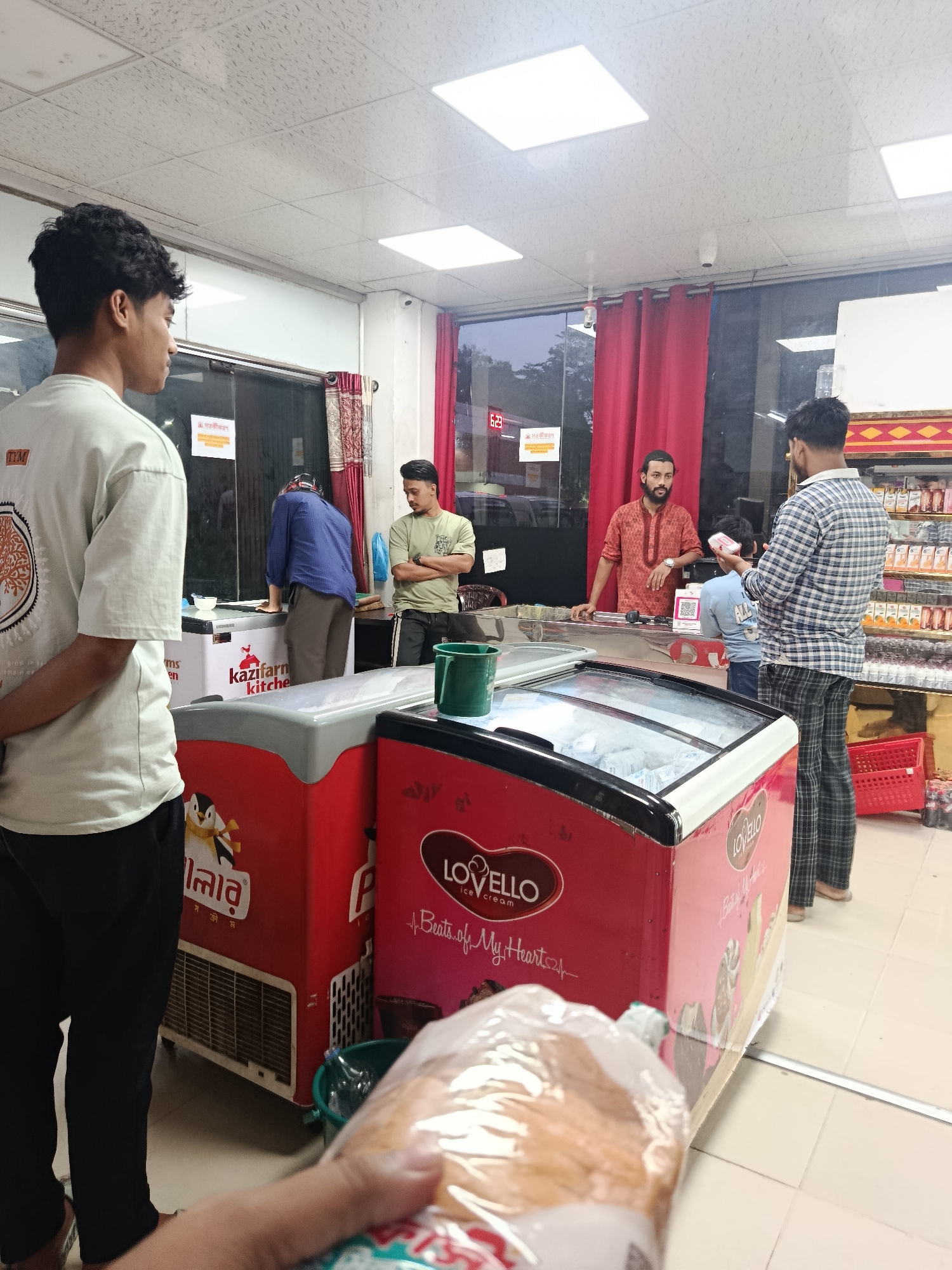 The image depicts the interior of a grocery store in Dhaka, Bangladesh. Several individuals are present: a young man is standing in the foreground, facing away from the camera. A man in a blue shirt is reaching into a freezer, while another man in a green shirt is standing with his arms crossed, observing. Behind the counter, a man is dressed in a traditional red shirt, possibly serving a customer who is holding a phone. A child also stands near the counter.
The store features ice cream freezers with brands like "Lovello" and "Kazifarm Kitchen". Red curtains are visible in the background. A sign reads "সতর্কীকরণ" (meaning "Warning" in Bengali). A clock on the wall reads "6:23". A bag of bread is held in the very foreground of the shot, obscuring parts of the scene.