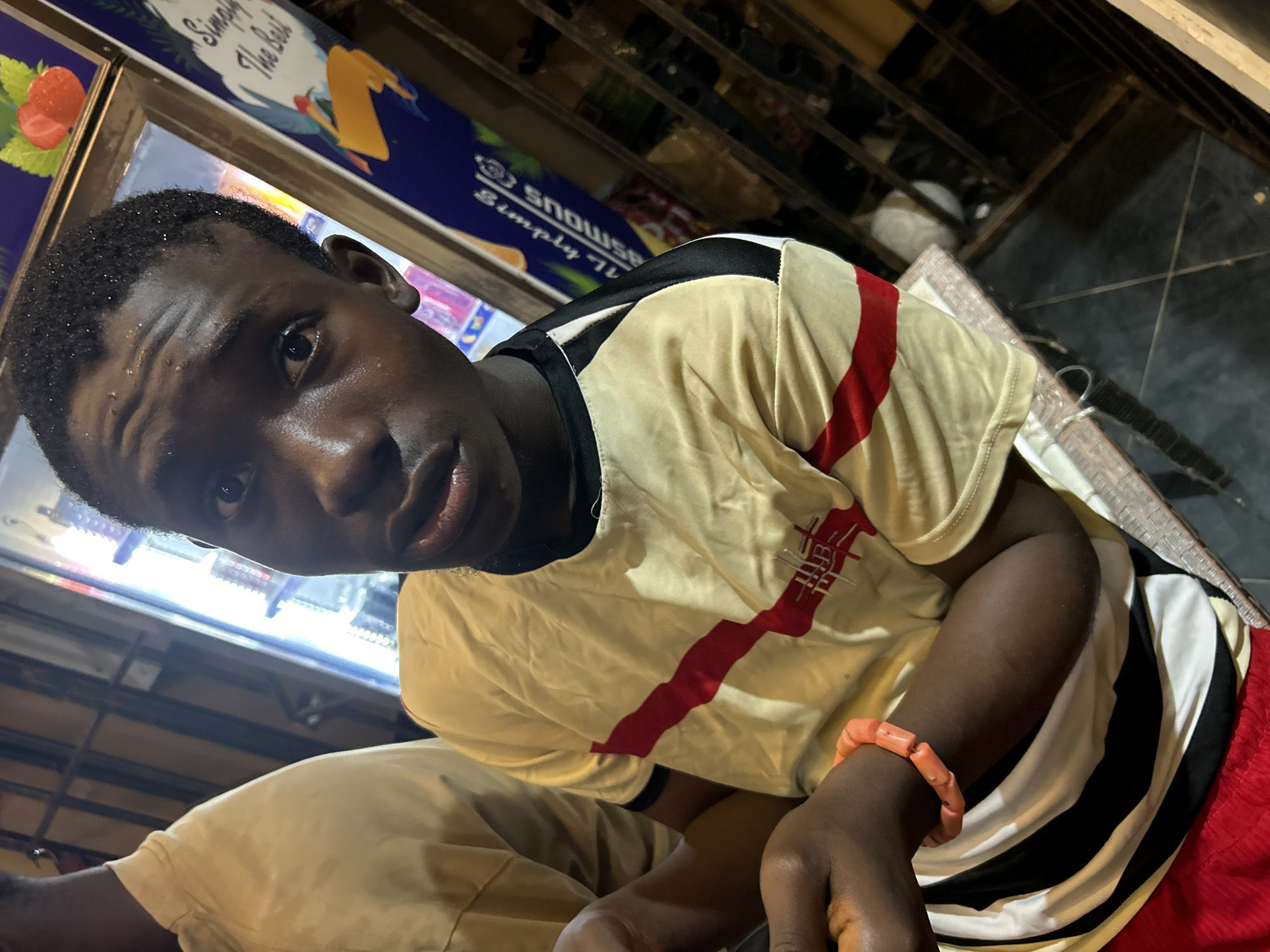 A medium shot captures a young Black male positioned to the right of the frame, looking directly at the viewer. He is seated at a table in what appears to be a food establishment, possibly a restaurant or bar in Ihiagwa, Nigeria. The background includes a refrigerated display case stocked with drinks, illuminated by bright lights, and a poster featuring strawberries and the text "Simply The Best." To the left of the main subject, another individual is partially visible, wearing a tan shirt. The main subject is wearing a yellow t-shirt with red and black stripes, a black trim collar, and a black and white striped pattern on his lower torso. On his left wrist, he wears a bright orange bracelet. His skin is shiny, suggesting sweat, and his hair is short and dark. The lighting in the scene is direct and somewhat harsh, creating reflections on his forehead and hair. The environment is dimly lit aside from the illuminated display case and direct lighting on the subjects.