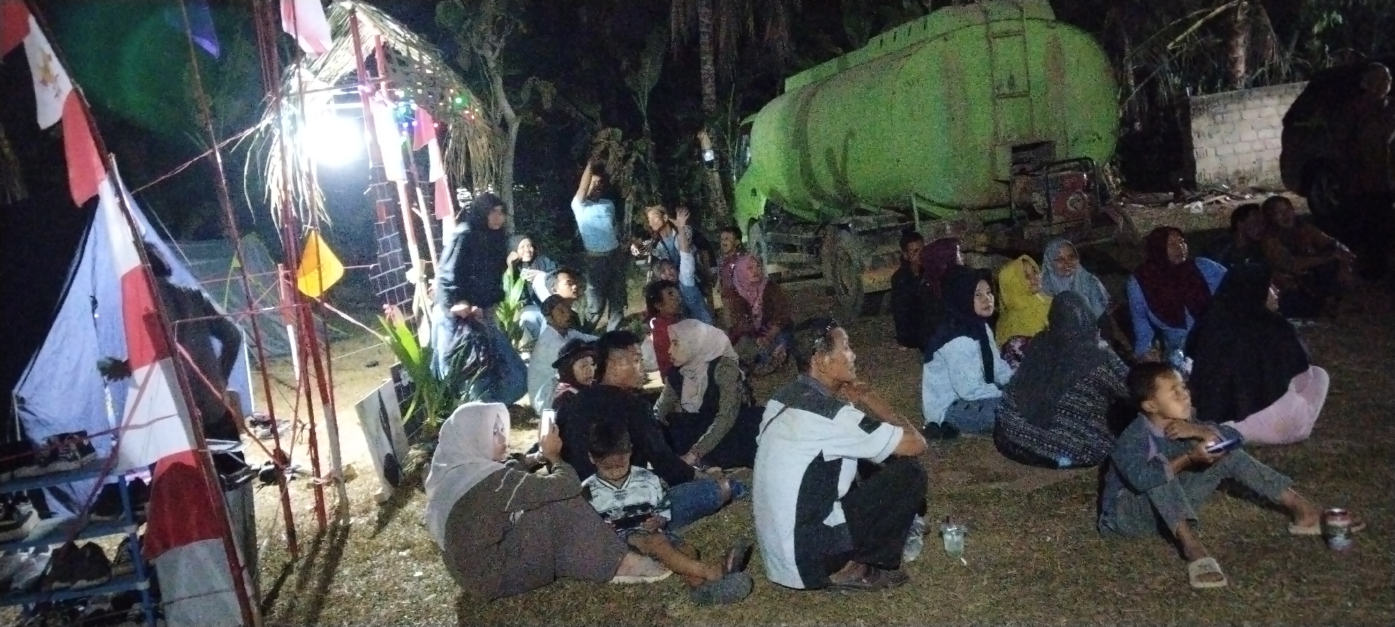 This image depicts an outdoor gathering at night in the city of Sido Rahayu, Indonesia. A group of approximately 20 individuals, of various ages and genders, are seated or standing on grassy ground. They are gathered near a makeshift structure adorned with flags and lights, and a large green water truck is visible in the background. Several people are looking towards something out of frame, with some individuals waving or raising their hands. The lighting suggests artificial illumination from the structure and possibly the truck. The environment appears to be rural or semi-rural, with palm trees and other vegetation visible. Some individuals are wearing headscarves, common in Indonesia.