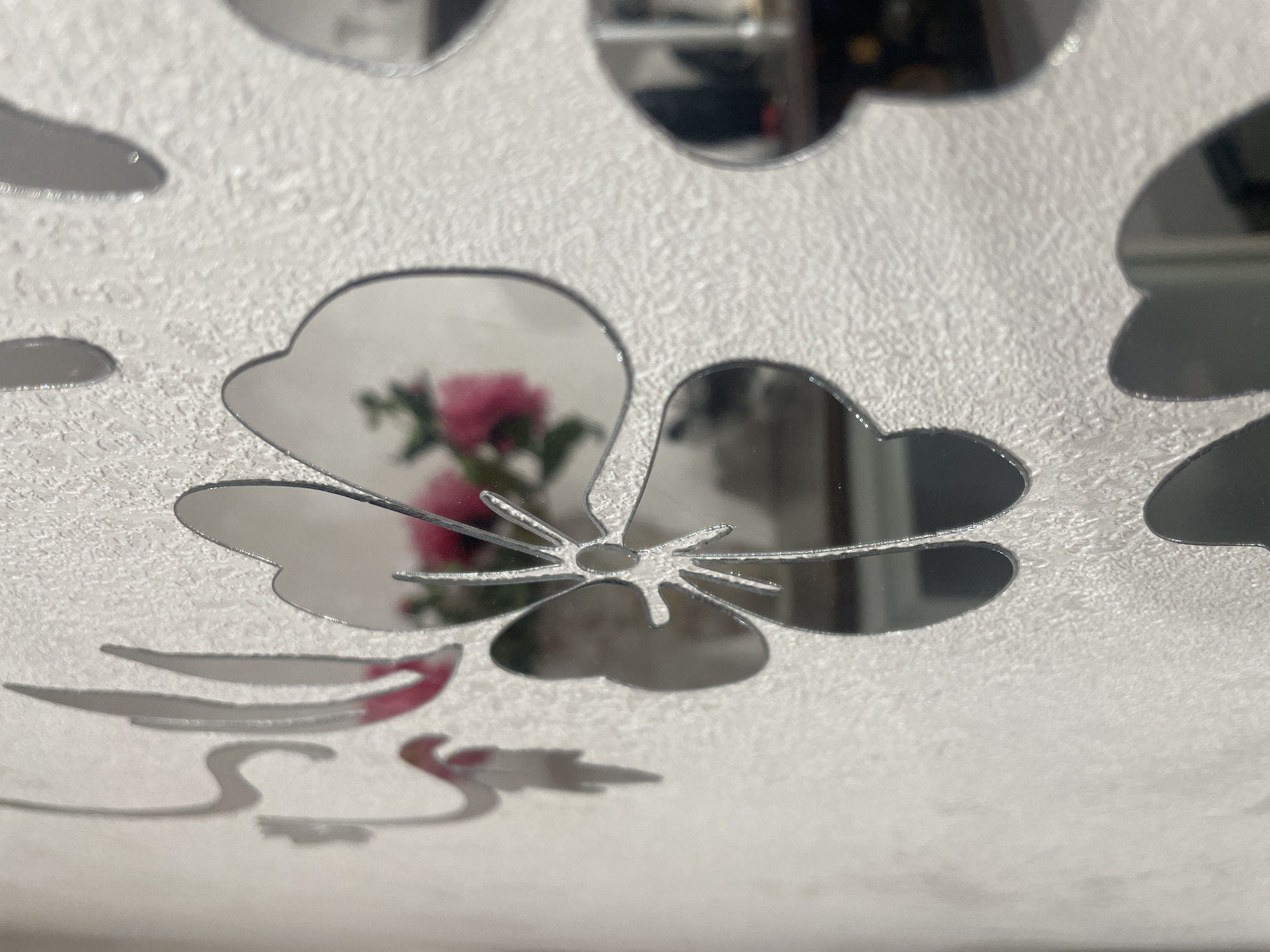 The image is a close-up, shallow depth-of-field shot of decorative mirrored flower stickers adhered to a textured, light-colored wall. The stickers are silver and have a reflective surface, showing distorted reflections of the surroundings. In the center, a large mirrored flower is prominent, with its petals and stamen clearly visible. A reflection of pink flowers and green leaves is visible within one of the petals. Other mirrored flower shapes are visible partially in the frame, some in focus and some blurred due to the shallow depth of field. The wall itself has a granular texture, possibly wallpaper or painted plaster. The lighting suggests an indoor environment, possibly a domestic setting. The location information "Aropakkuzi, Russia" is not visually supported by the image content.