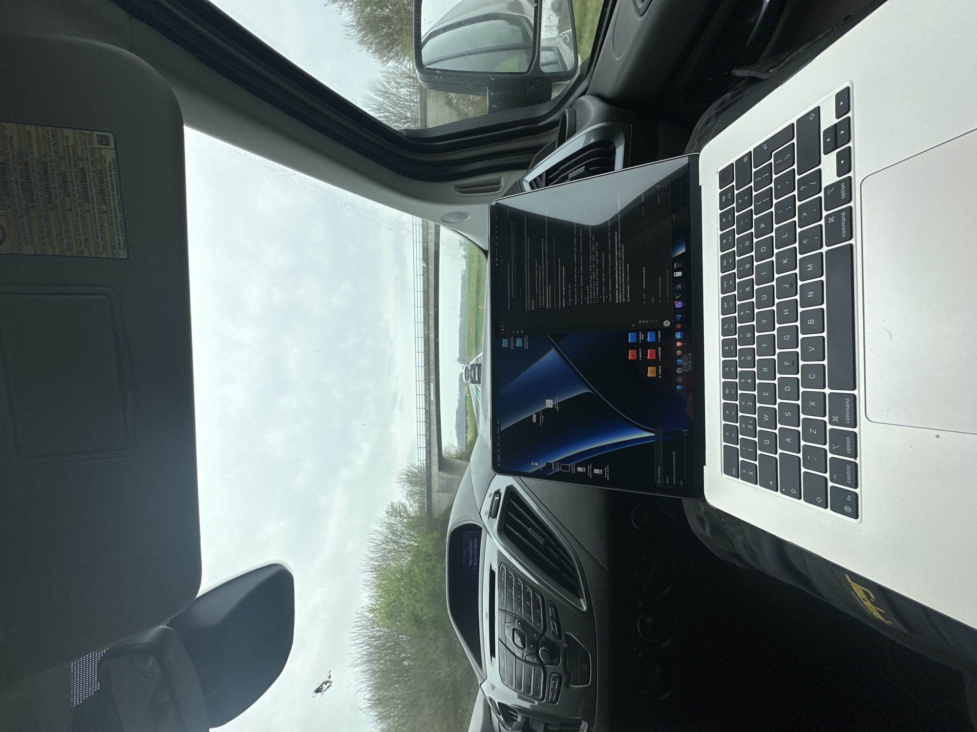 The image depicts an interior view from a vehicle, looking forward through the windshield towards a road and landscape in Druten, Netherlands. In the immediate foreground, a silver-colored MacBook laptop rests on a person's dark pants or lap. The laptop screen is illuminated, displaying a macOS desktop with a blue and black abstract wallpaper, a dock populated with numerous application icons, and two open windows. One window appears to be a document or text editor with content predominantly in Cyrillic script, while the other window's content is less discernible but appears to be a file browser or web page. An "iCloud" notification is visible near the bottom left of the screen.
The vehicle's dashboard is visible behind the laptop, featuring a dark gray plastic construction. The central console includes an audio unit with a small monochrome display indicating "07 audio," "Play music from," and "Bluetooth device." Below this are physical buttons and knobs for audio control, and to the right, vertical air vents. A rearview mirror hangs from the ceiling, and the passenger-side sun visor is visible above it, bearing a multi-language warning sticker, including the Dutch word "WAARSCHUWING" (warning). A small crack or defect is present on the upper left of the windshield. The passenger-side door panel and a portion of the side-view mirror, reflecting the vehicle's side, are visible on the right.
The exterior environment, observed through the windshield, shows a two-lane road stretching into the distance under an overcast sky. Green trees and brush line the sides of the road. A multi-lane concrete bridge spans the road in the mid-distance. Another vehicle is visible in the distance on the road, traveling in the same direction. No individuals are directly visible, though a person is implied to be operating the laptop and holding the camera within the vehicle.