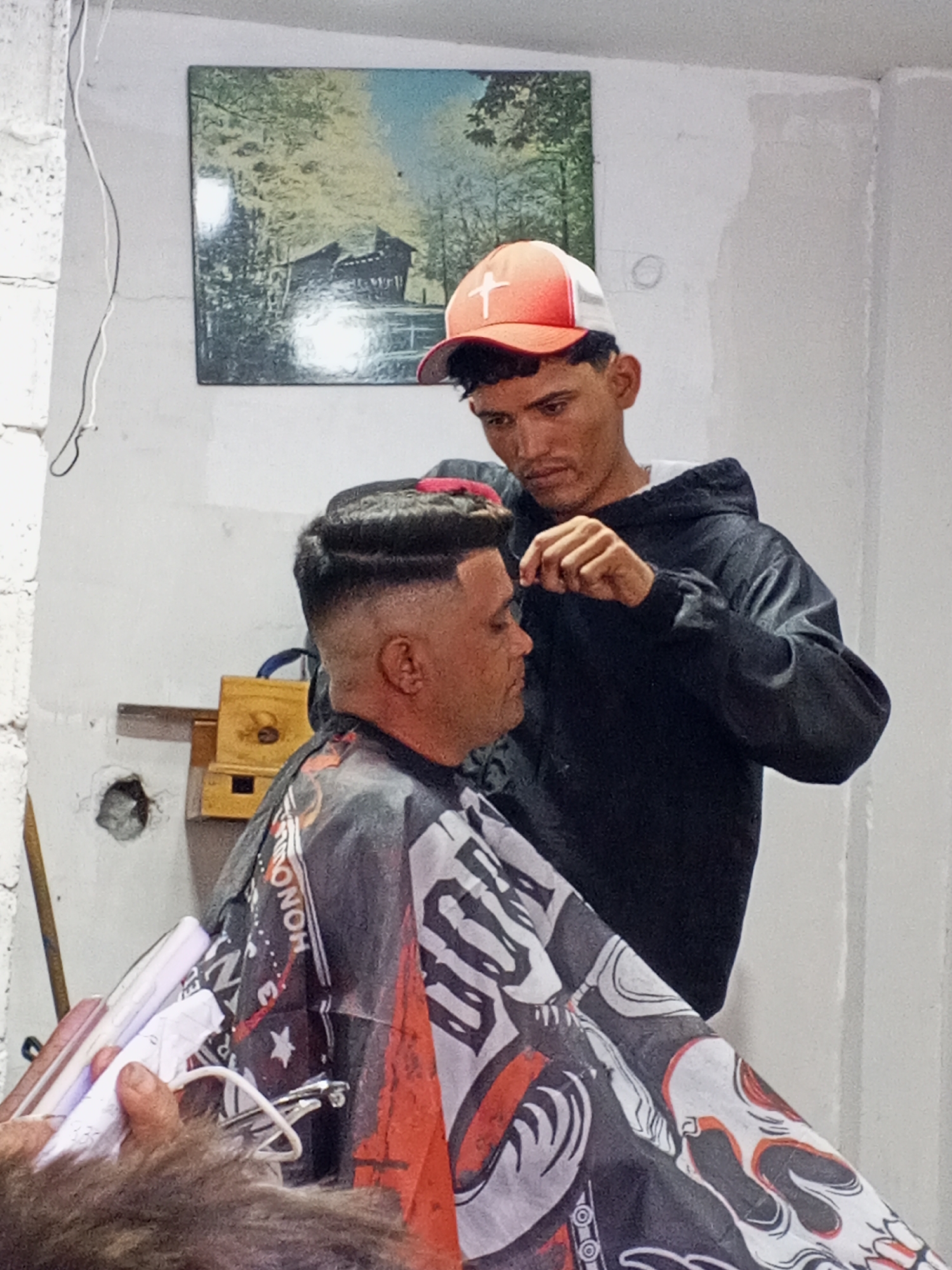 In an indoor setting, a barber with a red and white baseball cap, wearing a black hoodie, is giving a haircut to a seated customer. The customer is draped with a black, white, and red patterned barber's cape. A picture hangs on the wall behind them. Various barber tools are visible in the foreground. The location is in the city of Unknown, Unknown.