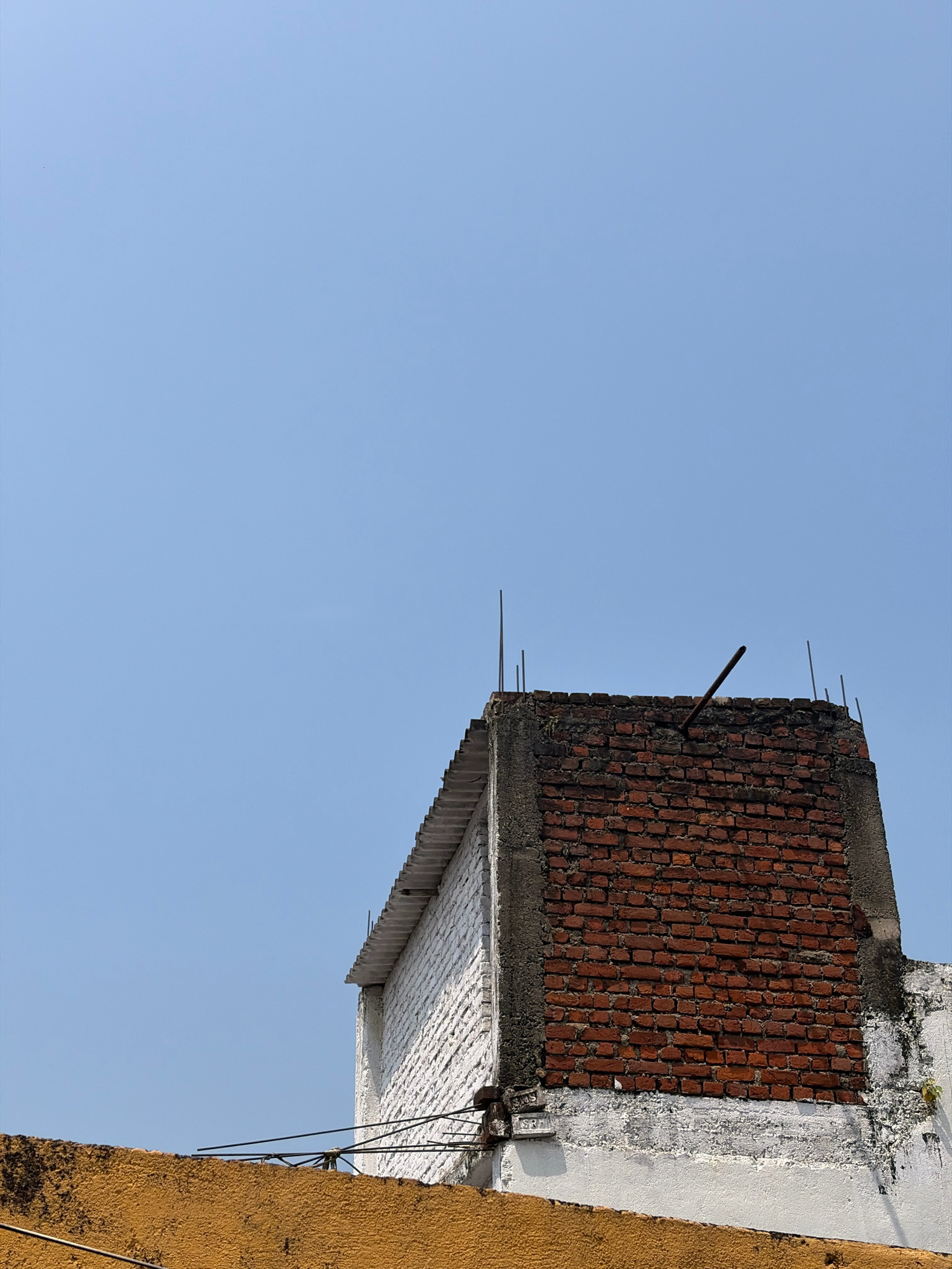 The image shows a close-up of a rooftop in Jamshedpur, India, against a clear blue sky. The rooftop features a red brick chimney structure with rough concrete edges. Several metal rods protrude from the top of the chimney, and one is bent at an angle. To the left of the chimney, a white-painted structure with a corrugated roofline is visible. The foreground is dominated by an orange-colored wall, partially obscuring wires and some of the white structure below. The scene suggests an urban environment, likely residential, with exposed building materials and utility wires.