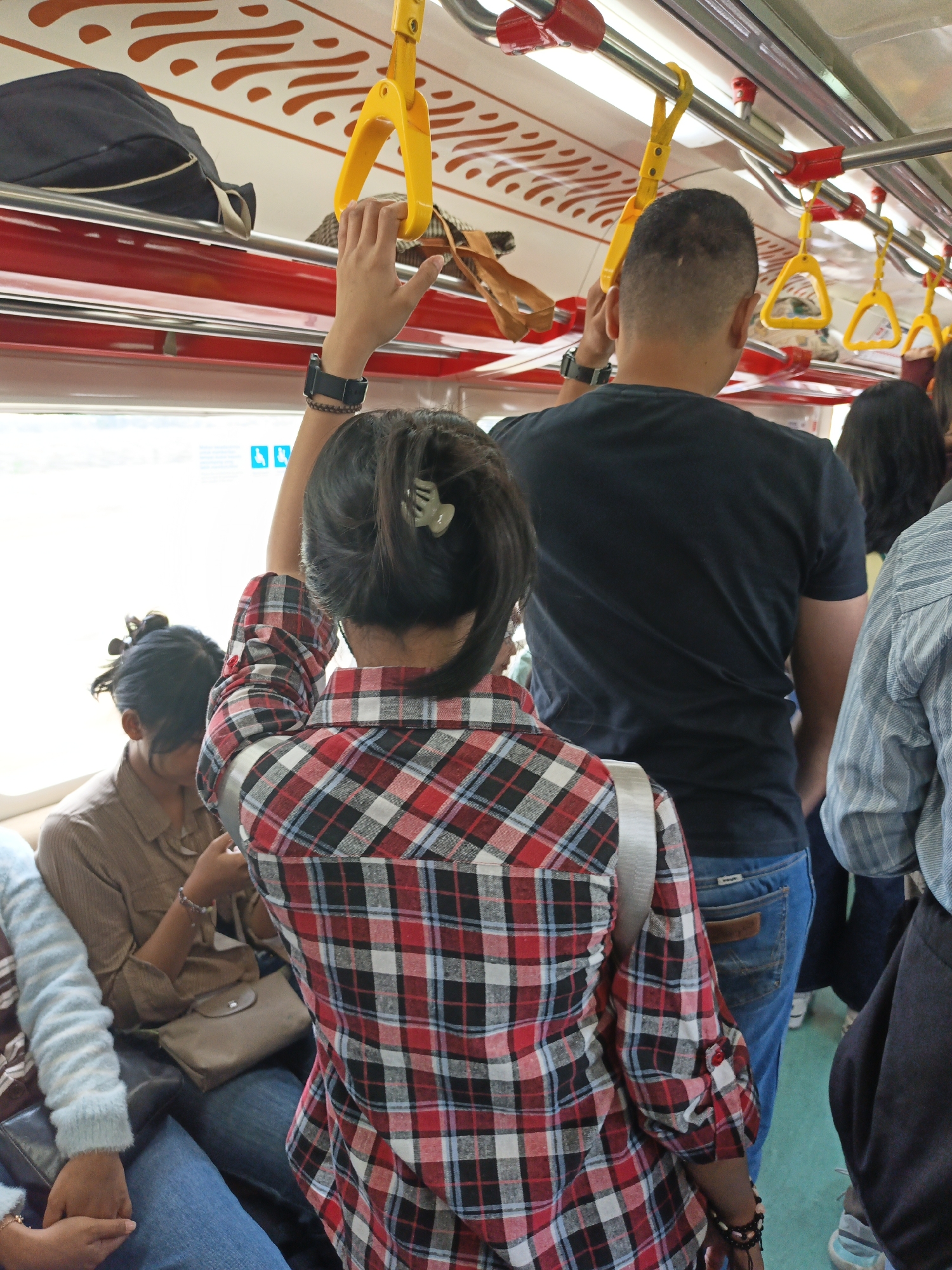 This image captures the interior of a public transportation vehicle, likely a train or bus, filled with passengers. A woman in the foreground is holding onto a yellow handrail, her back to the camera. She is wearing a red and black plaid shirt and has a light green hair clip in her dark hair. To her right, a man wearing a black t-shirt is also holding a yellow handrail. Several other passengers are visible in the background, some seated and some standing. The vehicle's interior features red and white seating and metal handrails. The image appears to have been taken in Pokak, Indonesia, suggesting this is a common mode of transport in the area.