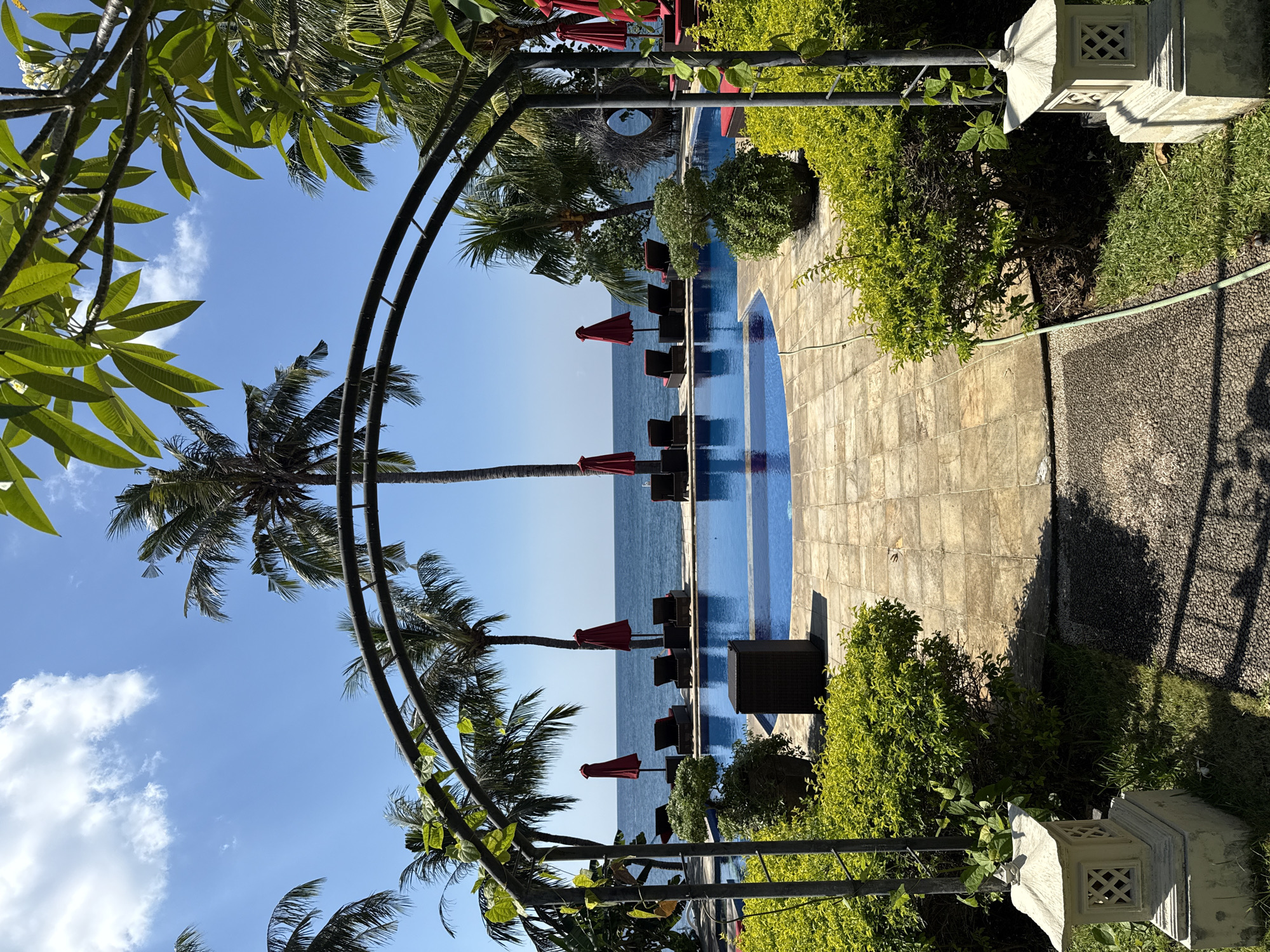 This is a medium shot, eye-level, vertical composition of an outdoor swimming pool area with the ocean in the background, likely in Kubu, Indonesia. The foreground is dominated by a dark metal archway with leafy vines. To the left of the archway, a stone lantern sits on the grass. A pathway made of small grey stones leads towards the arch. The swimming pool is a vibrant blue and is surrounded by light brown square tiles. A few dark wicker chairs with red cushions are visible on the tiled area.
Beyond the pool, a line of dark lounge chairs with red umbrellas are arranged along the edge of the pool, facing the ocean. The ocean is a calm, deep blue, meeting a clear blue sky with a few scattered white clouds. Several palm trees, characteristic of tropical locations, are visible throughout the scene, with lush green foliage. The overall impression is of a serene and inviting resort setting. There are no people visible in the image.