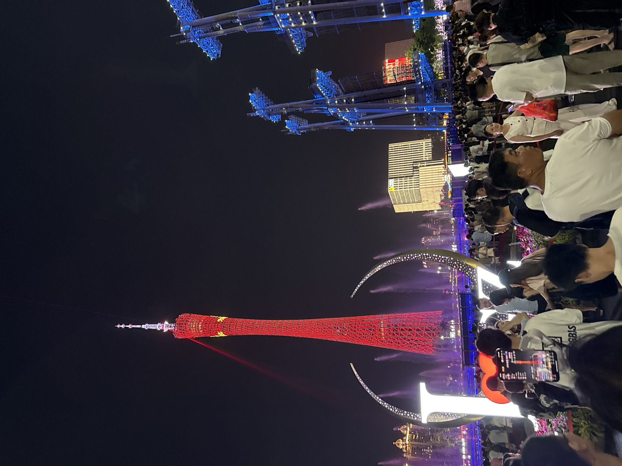 This image depicts a nighttime scene in Tianhe District, China, likely during a public event. The primary subjects are a large crowd of people gathered to watch a spectacle. In the background, a tall, red lattice tower, illuminated with yellow accents, dominates the skyline, possibly a landmark like the Canton Tower. Overhead, illuminated blue structures, resembling bridges or elevated walkways, are visible against the dark sky, also adorned with lights. A modern, striped building is present in the mid-ground. The foreground is filled with people, many holding up phones to capture the event, suggesting a performance or display. Some individuals are dressed casually, while others appear to be enjoying a festive atmosphere. The lighting suggests it's nighttime, and the presence of the crowd and illuminated structures points towards an organized event, perhaps a light show or festival. The overall mood appears to be one of engagement and enjoyment. Some visible text on a person's t-shirt appears to read "GBIS".