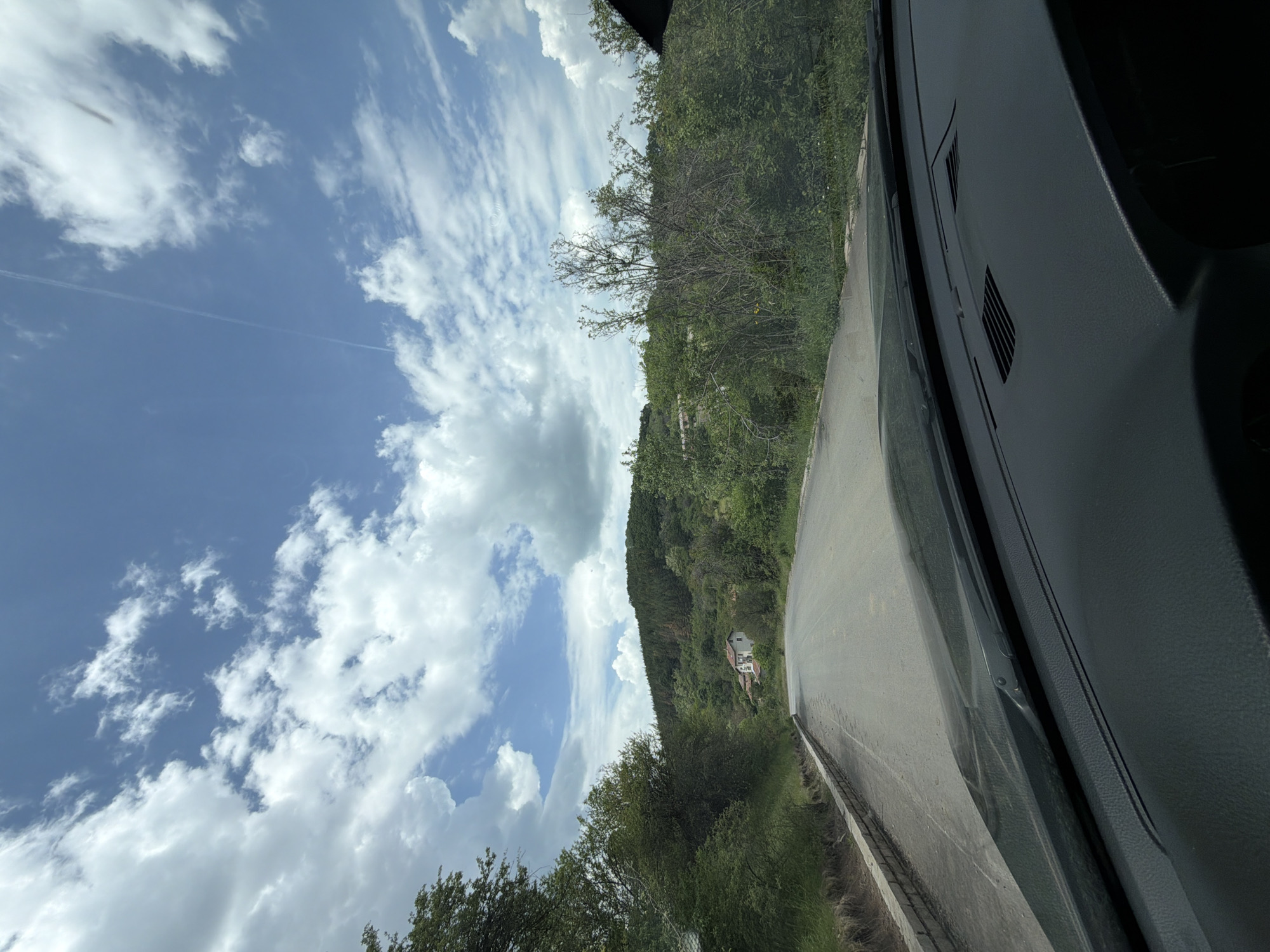 A view from inside a vehicle traveling on a paved road. Lush greenery covers the hillside to the right of the road, while a partly cloudy sky dominates the left portion of the frame. Buildings are visible amidst the trees on the hillside. The interior of the vehicle is visible in the right side of the image, featuring the dashboard. The location is Gurgulyat, Bulgaria.