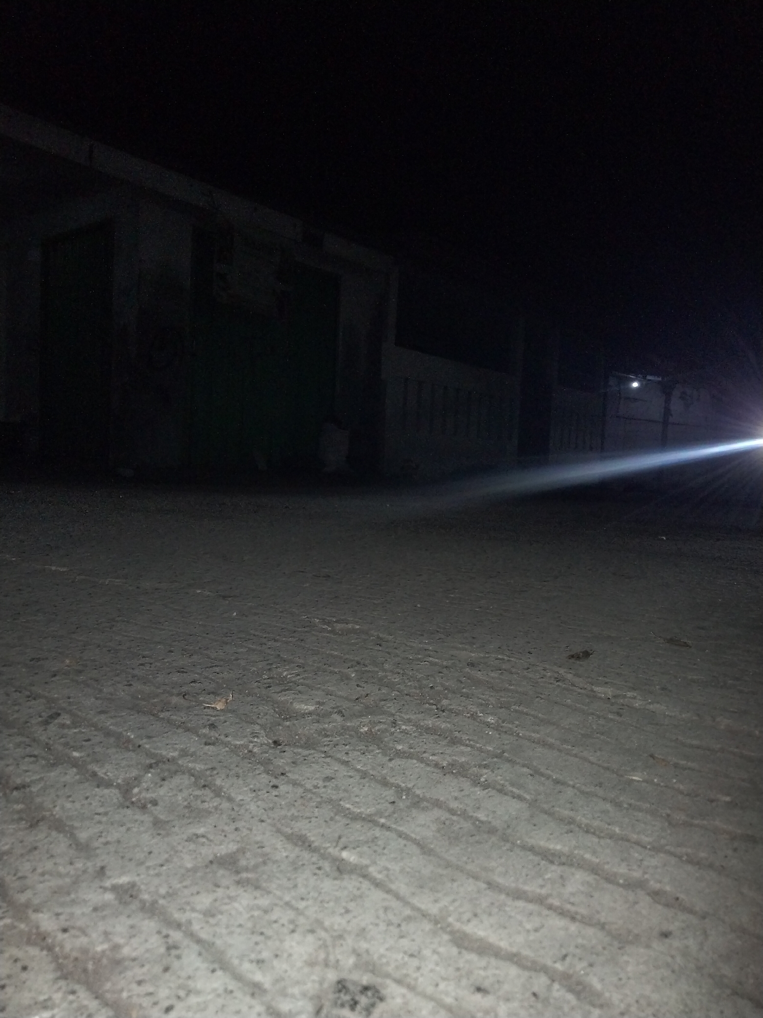 The image captures a dimly lit outdoor scene at night. A paved surface, likely a street or pathway, occupies the foreground, showing some texture and debris. In the background, a dark building with visible architectural features such as walls and what appear to be doorways or openings is present. A bright light source on the right side of the image casts a strong beam across the scene, illuminating a portion of the building and the air. The overall impression is one of an urban or semi-urban environment at night, possibly in a less developed area due to the rustic appearance of the building and the paving. The location is specified as Cariu, Indonesia.