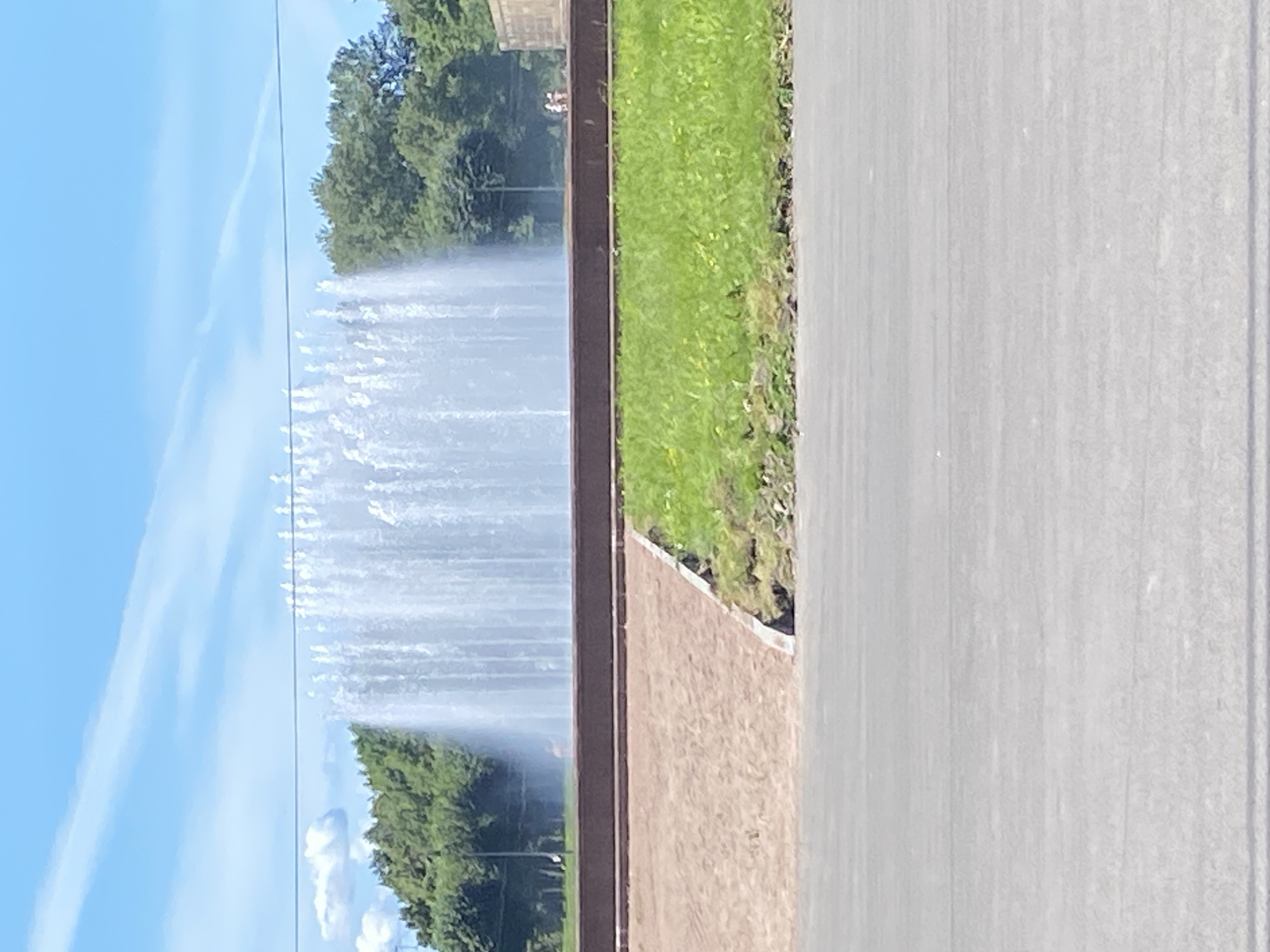 The image captures a wide, tall, multi-tiered fountain spraying water upwards against a backdrop of green trees and a bright blue sky with scattered white clouds. In the foreground, a paved area transitions into a strip of brown sand and then a vibrant green lawn. The fountain appears to be in a park or public space within Saint Petersburg, Russia. Two small figures, possibly people, are visible in the distance near a stone structure to the right of the fountain. The overall scene suggests a sunny day in an urban recreational area.