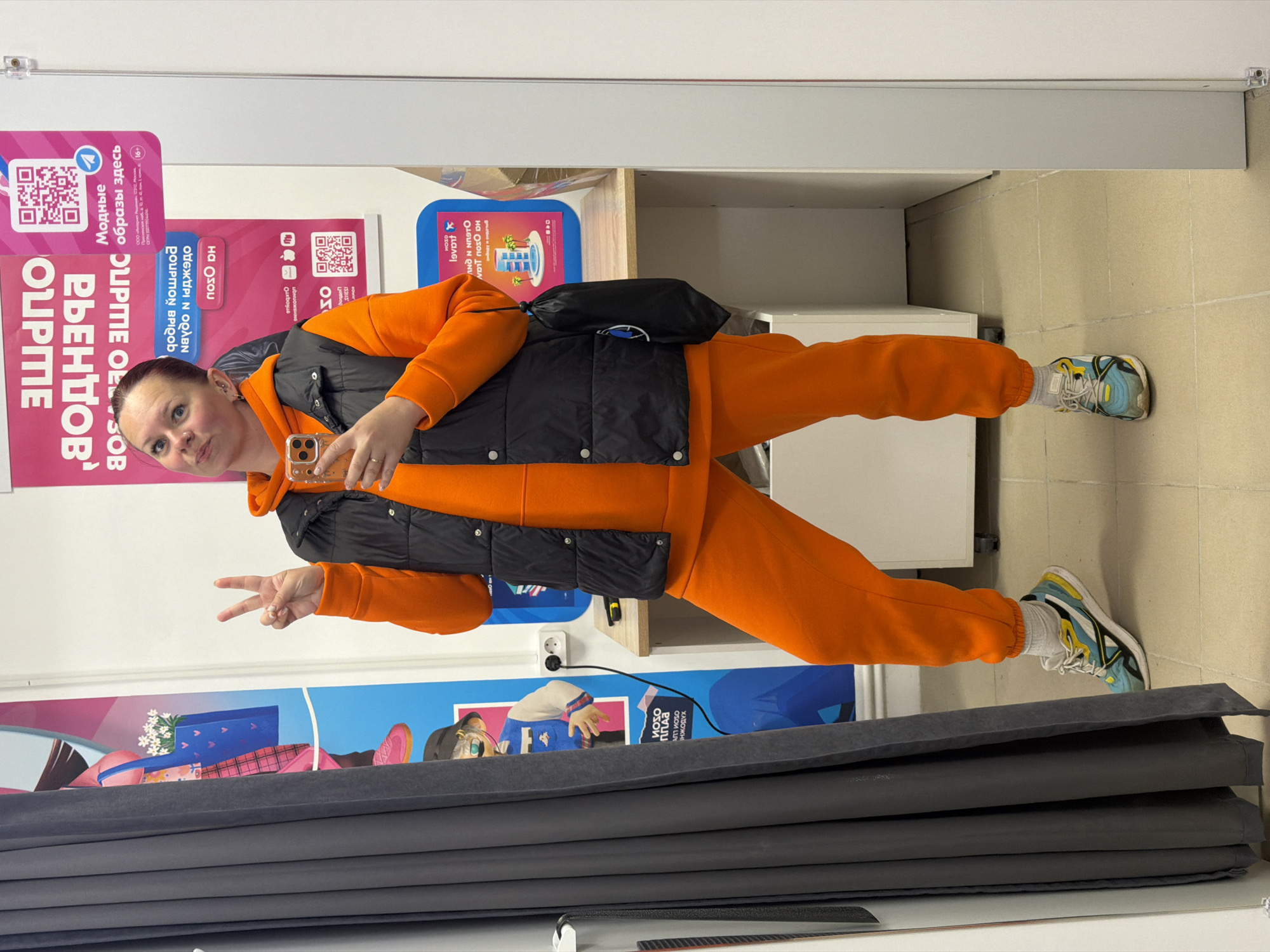 A woman stands at an angle, giving a peace sign while holding a phone in her right hand, wearing an orange hoodie, orange sweatpants, and a black vest, with blue and yellow sneakers. She is in what seems to be a retail or commercial space. The location is the city of Chaikovsky, Russia, based on the Cyrillic script visible on the posters behind her that include the city's name. Behind her are advertisements and a wall display. In the foreground, there are what look like folded grey upholstery materials. The floor is tiled.