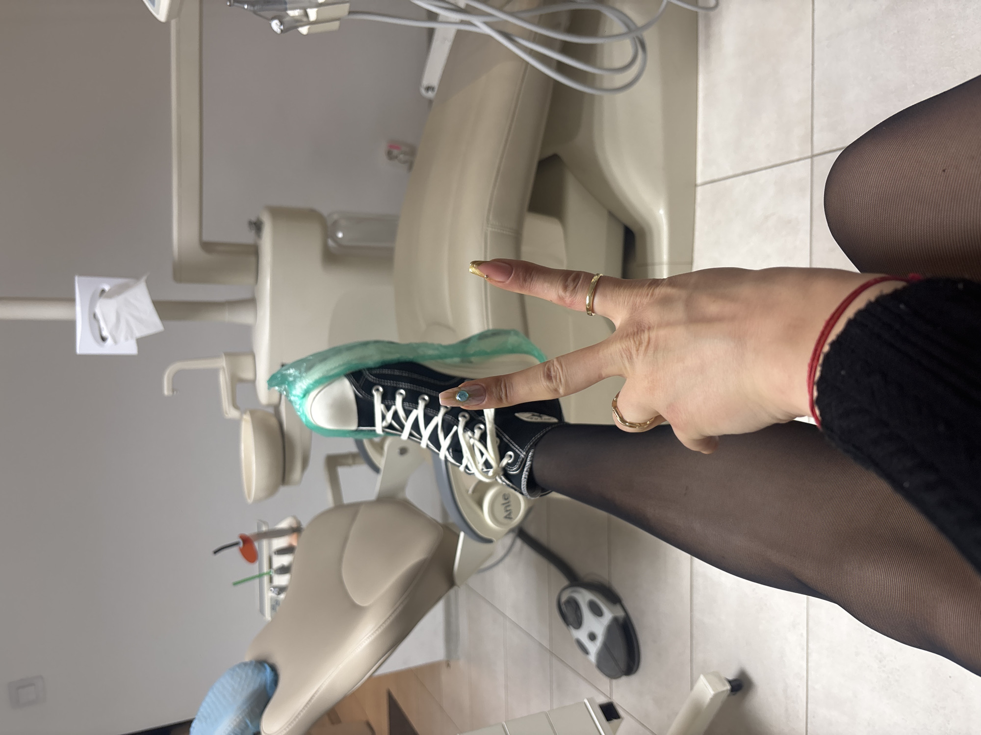 The image shows a person sitting in a dental chair, wearing black tights and a black sweater. Their right leg is extended with a black and white high-top sneaker on their foot. The sneaker has a light green sole and white laces. The person's right hand is making a peace sign over the sneaker. They have gold-painted fingernails and are wearing rings on two fingers.
The background consists of the dental chair, covered with a light blue disposable protector, and dental equipment, including a light fixture and tubes. The location appears to be a dental office. The city and country are not discernable.