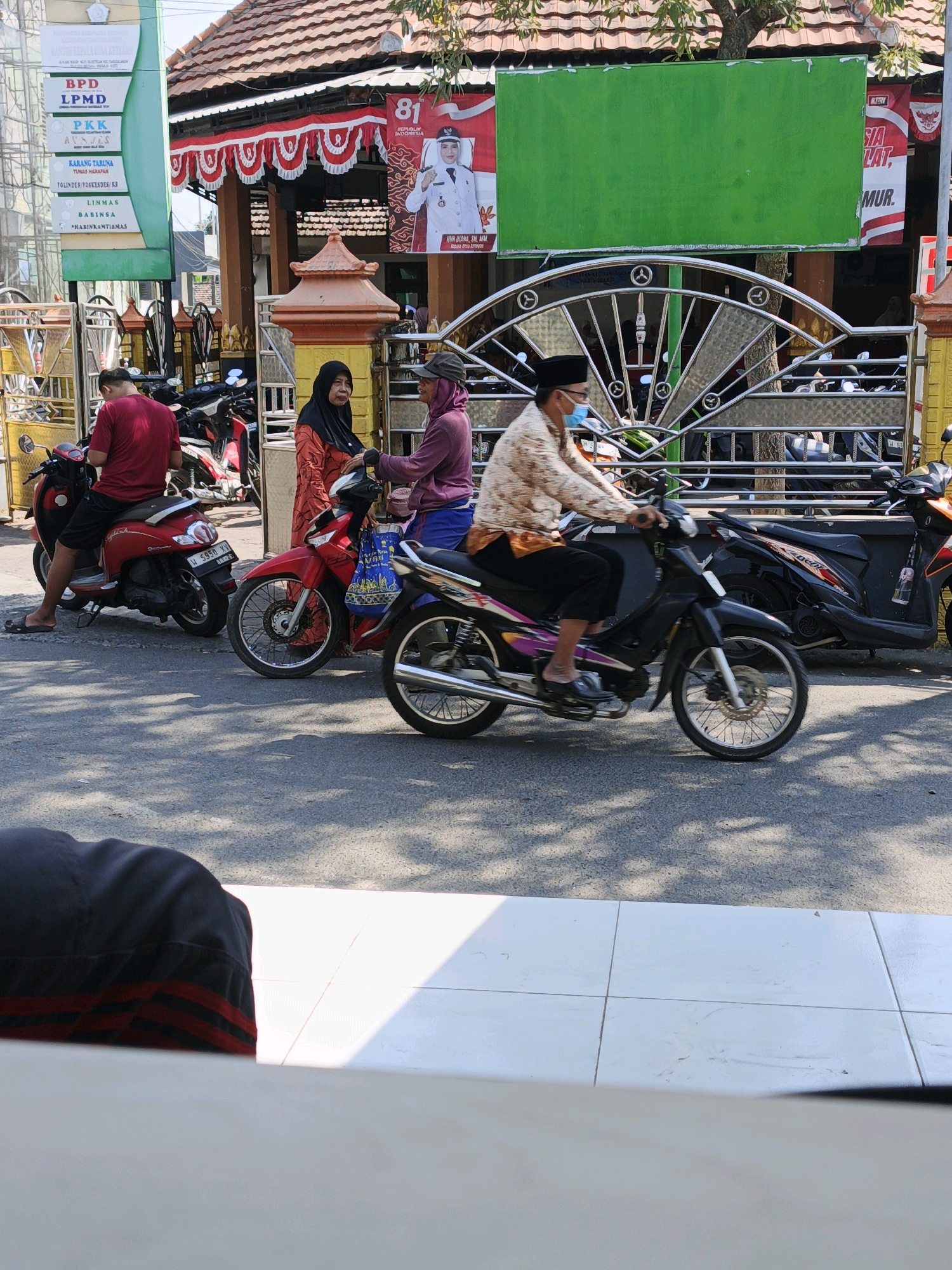 In Ketegan, Indonesia, an outdoor scene unfolds with parked motorcycles and several individuals. On the left, a person in a red shirt and black shorts sits on a red scooter, facing right. To their right, a woman in an orange and black patterned dress and headscarf stands near a red motorcycle. A man in a patterned shirt, black pants, black peci, and blue mask rides a black motorcycle from left to right across the frame, passing through a decorative metal gate. Behind the gate, more motorcycles are parked. The background features a building with a tiled roof and red and white banners, including one with the number "81" and an image of a woman in uniform. A green billboard is mounted above the gate. The ground is asphalt, with dappled sunlight and shadows from trees overhead. In the foreground, a person's lap covered by a dark blue and red striped garment is visible, with white tiles below.