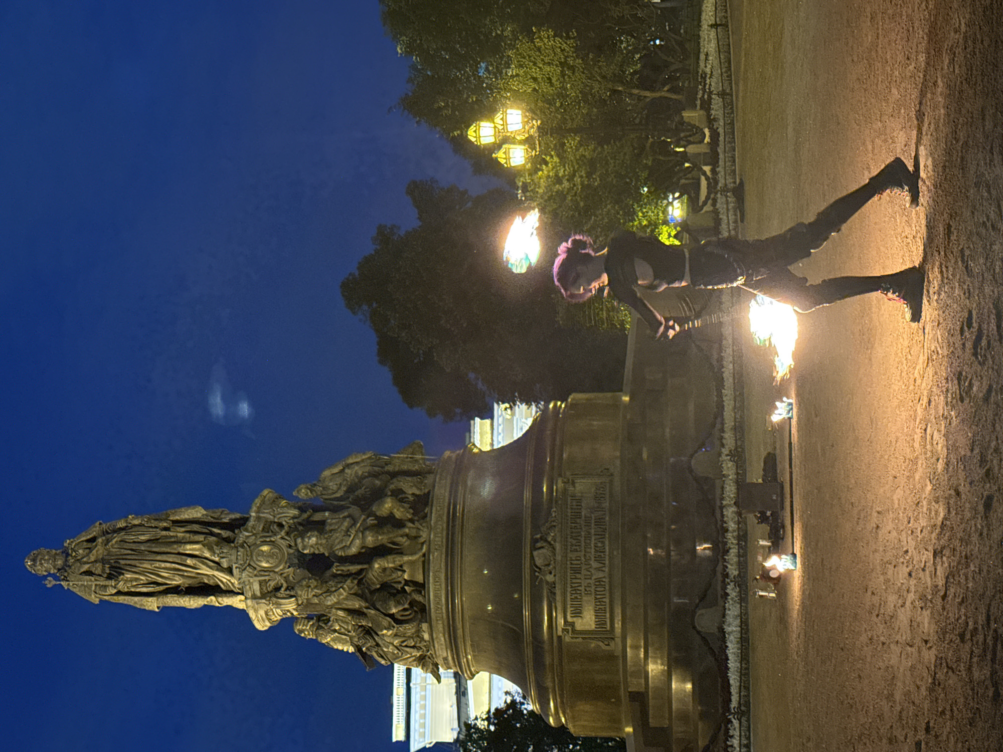 A fire performer with purple-dyed hair is performing in front of the Monument to Catherine the Great at night in Palace Square, Saint Petersburg, Russia. The monument, a large bronze sculpture of Catherine the Great standing atop a pedestal surrounded by allegorical figures, is illuminated by spotlights. The performer, dressed in dark, contemporary clothing, is twirling a flaming prop, creating a vibrant spectacle against the darkening sky. Several other smaller flames are visible on the ground around the performer, possibly part of the performance or setting. In the background, trees and ornate streetlights of Palace Square are visible, with the illuminated facade of the General Staff Building also discernible. The ground is covered in sand and gravel.