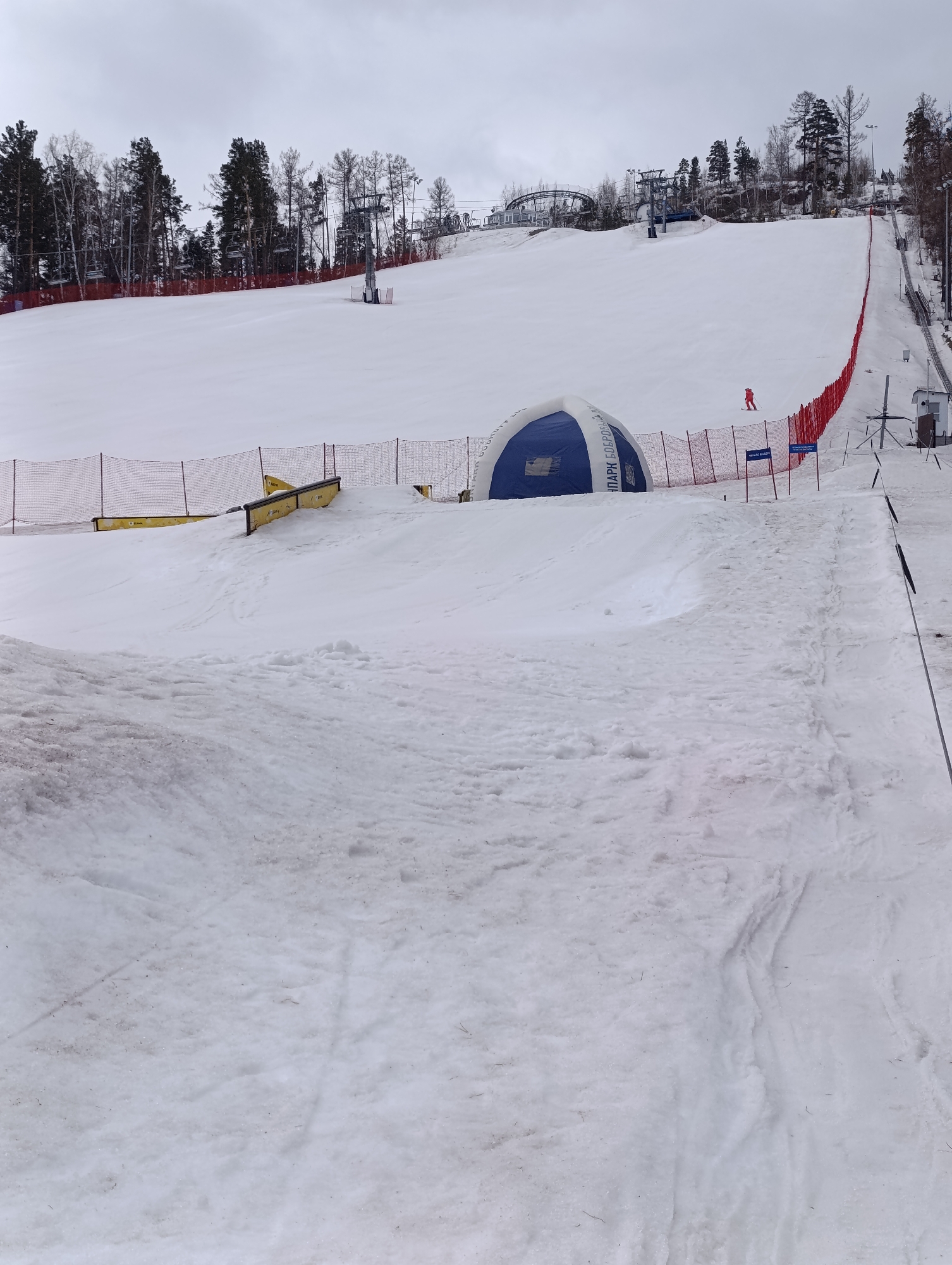 The image shows a snowy ski slope on a cloudy day in what is likely the Bobrovy Log Fanpark in Krasnoyarsk, Russia. The foreground features uneven snow, showing signs of melting. In the midground, there's a blue and white inflatable structure enclosed by orange safety netting, and a single person in red can be seen skiing down the slope. Yellow rails are also present, possibly for snowboarders. The background displays ski lifts, trees, and more snow covering the slope.