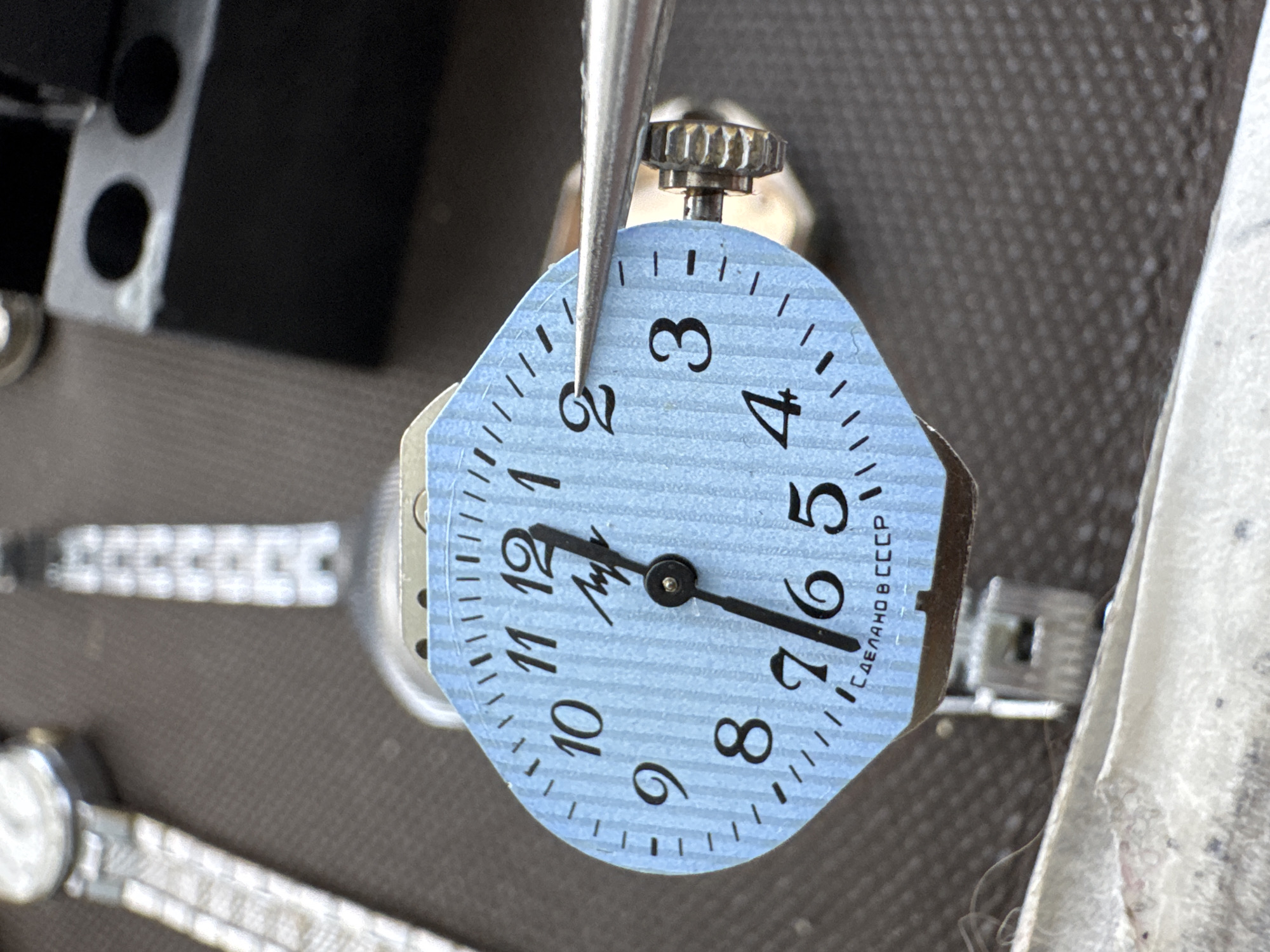 A close-up, high-angle shot shows a light blue watch dial with black numbers and markings. The dial has a subtle vertical stripe pattern. At the center of the dial, black hour and minute hands are visible. The hour hand points slightly past the 12, and the minute hand points to the 7. The words "СДЕЛАНО В СССР" (Made in USSR) are printed in small black letters at the bottom of the dial. A pair of silver tweezers is positioned to touch the number 2 on the dial. In the background, parts of other watches and watch components are visible, including a watch band on the left and a black plastic component on the right. The overall setting appears to be a workbench or repair area. There is no indication of the city of Kempelevo, Russia in the image.