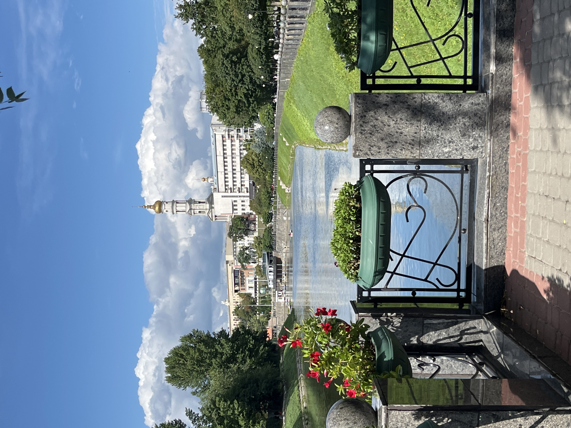 This is an eye-level, medium shot of a riverfront scene in Kharkiv, Ukraine, likely on a sunny day with scattered clouds. In the foreground, a granite balustrade with ornate black metal railings separates a paved walkway from the river. Dark green planters sit atop the balustrade, one containing vibrant red flowers and lush green foliage, and another filled with green plants. The river, reflecting the blue sky and white clouds, flows towards the left of the frame. On the far bank, a grassy embankment is lined with trees and a decorative railing. Beyond the trees, buildings are visible, including a prominent white structure with a golden-domed tower, characteristic of Orthodox churches in the region. Several boats, including a ferry and smaller vessels, are on the river. People are visible walking along the embankment on the far side of the river. The overall impression is a scenic and tranquil urban landscape.