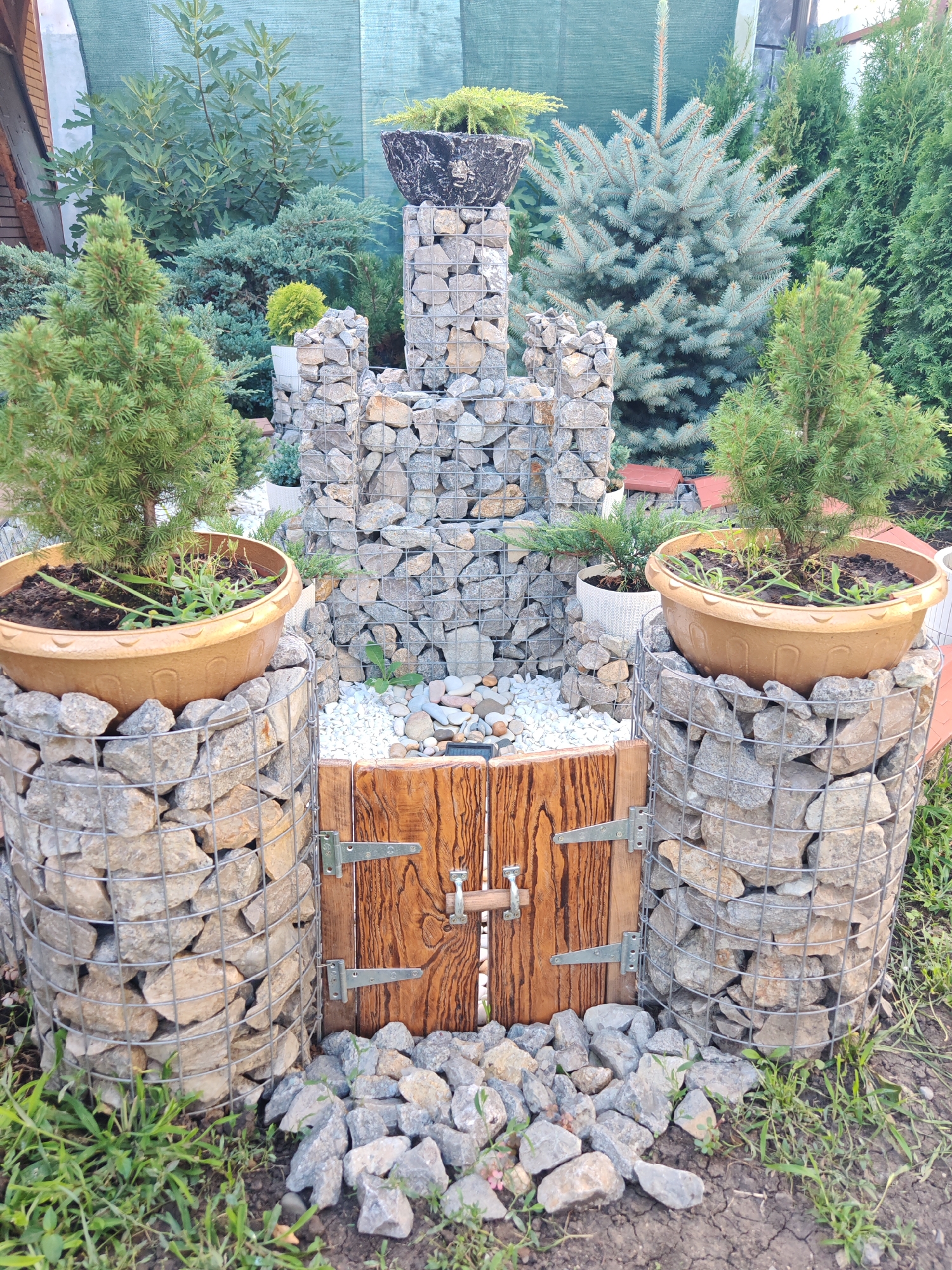 The image depicts a garden feature resembling a castle, constructed from gabions (wire cages filled with stones). Two cylindrical gabions, each topped with a potted plant, flank a central wooden door. The central structure is also made of gabions, forming ramparts and towers. The scene is set in a garden with various coniferous trees and shrubs, and the ground is covered with grass and stones. The location is identified as Oktyabrskiy, Russia. No people are visible in the image.