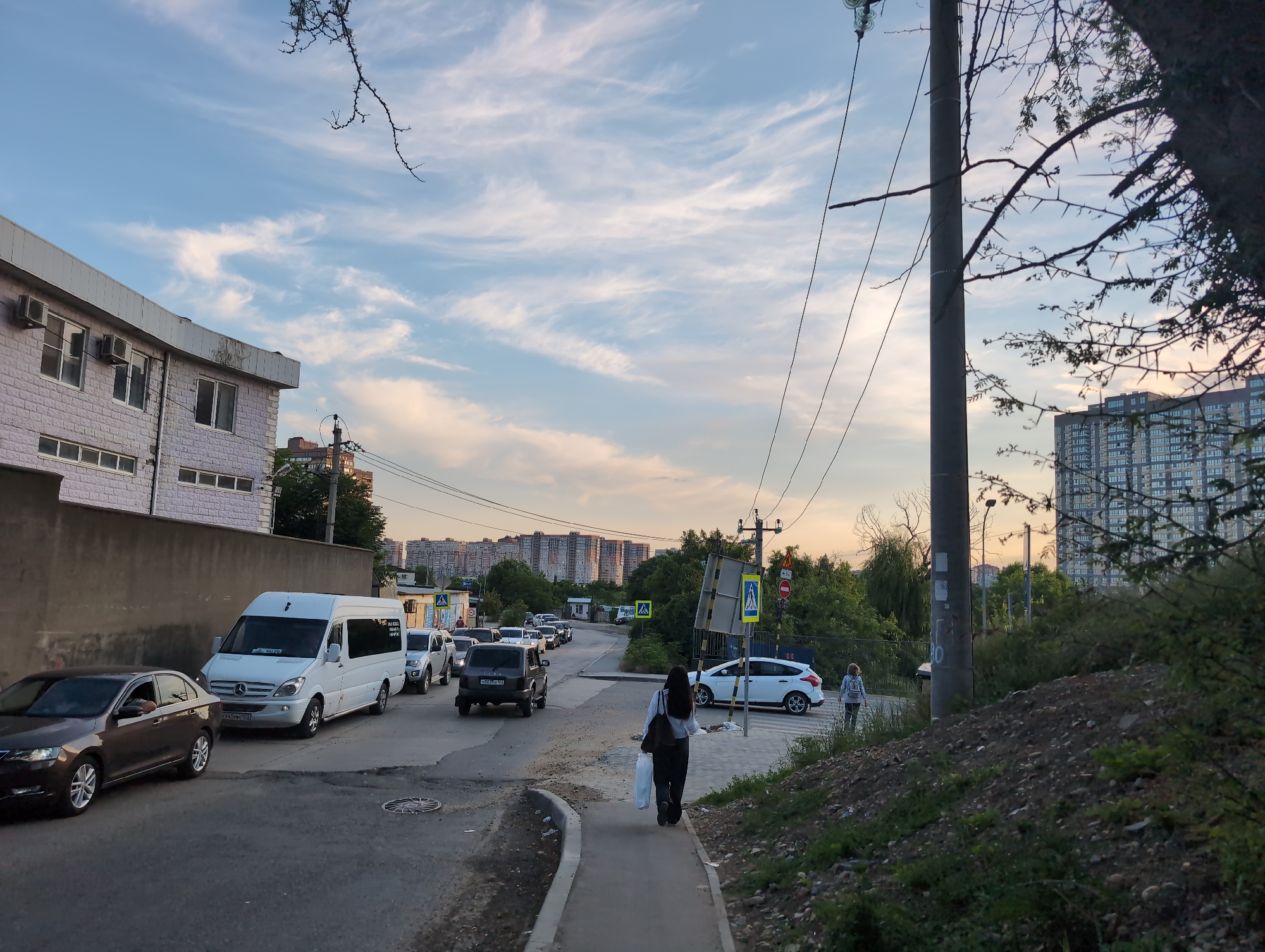 The image captures a street scene in Krasnodar, Russia, during the late afternoon or early evening, indicated by the warm lighting and the soft hues in the sky. The perspective is from a street level looking towards a road with moderate vehicular traffic and pedestrians.
On the left side of the road, a light-colored, multi-story building with a light grey facade and visible air conditioning units lines the street. Adjacent to it, a dark grey concrete wall runs along the sidewalk. Several cars are parked or moving on the road. A dark brown sedan is in the foreground, followed by a white Mercedes-Benz van, a silver SUV, and other vehicles further down the road.
A paved sidewalk runs along the right side of the road. Two individuals are walking on this sidewalk. The person in the foreground is wearing dark clothing and carrying two white bags. Further down the sidewalk, another person, possibly a woman with blonde hair, is walking away from the camera.
The road itself appears to be a two-lane street with some wear and tear, including visible patches and cracks in the asphalt. Pedestrian crossings are marked with white stripes, and traffic signs, including a pedestrian crossing sign and a prohibition sign, are visible.
In the background, a densely populated residential area with numerous tall apartment buildings is visible under a sky filled with wispy, white clouds against a pale blue backdrop. The overall environment suggests an urban setting, likely a residential neighborhood within Krasnodar. The presence of utility poles and electrical wires indicates infrastructure typical of a city. Lush green trees and shrubs are scattered throughout the scene, particularly on the right side of the road, suggesting some natural landscaping amidst the urban development.