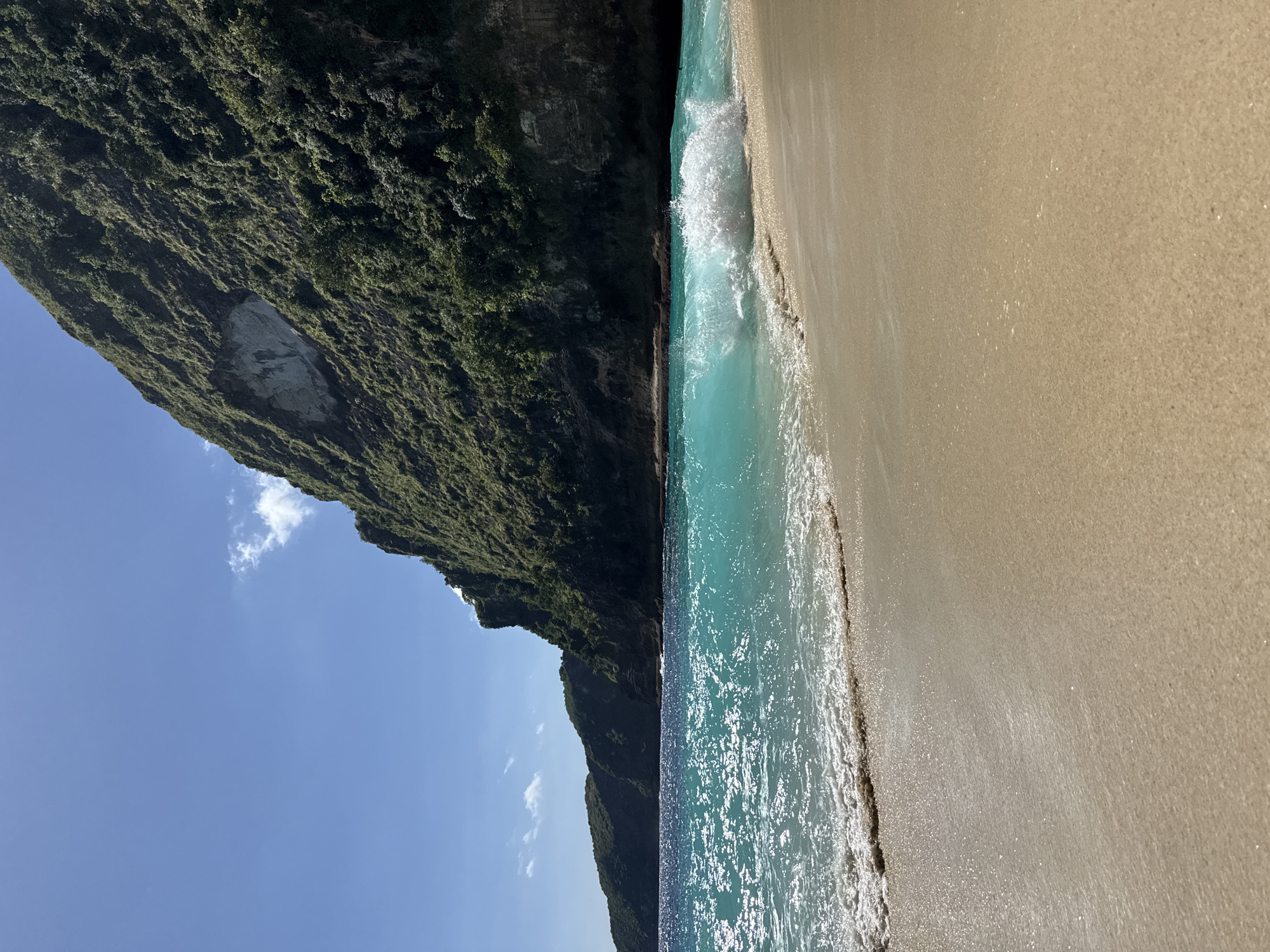The image shows a beach scene in Bunga Mekar, Indonesia. A cliff covered in dense green vegetation dominates the left side of the image. The right side displays a sandy beach meeting the turquoise water of the ocean. A small wave is breaking on the shore. The sky is visible in the upper left corner with a few small clouds. The image is rotated 90 degrees clockwise.