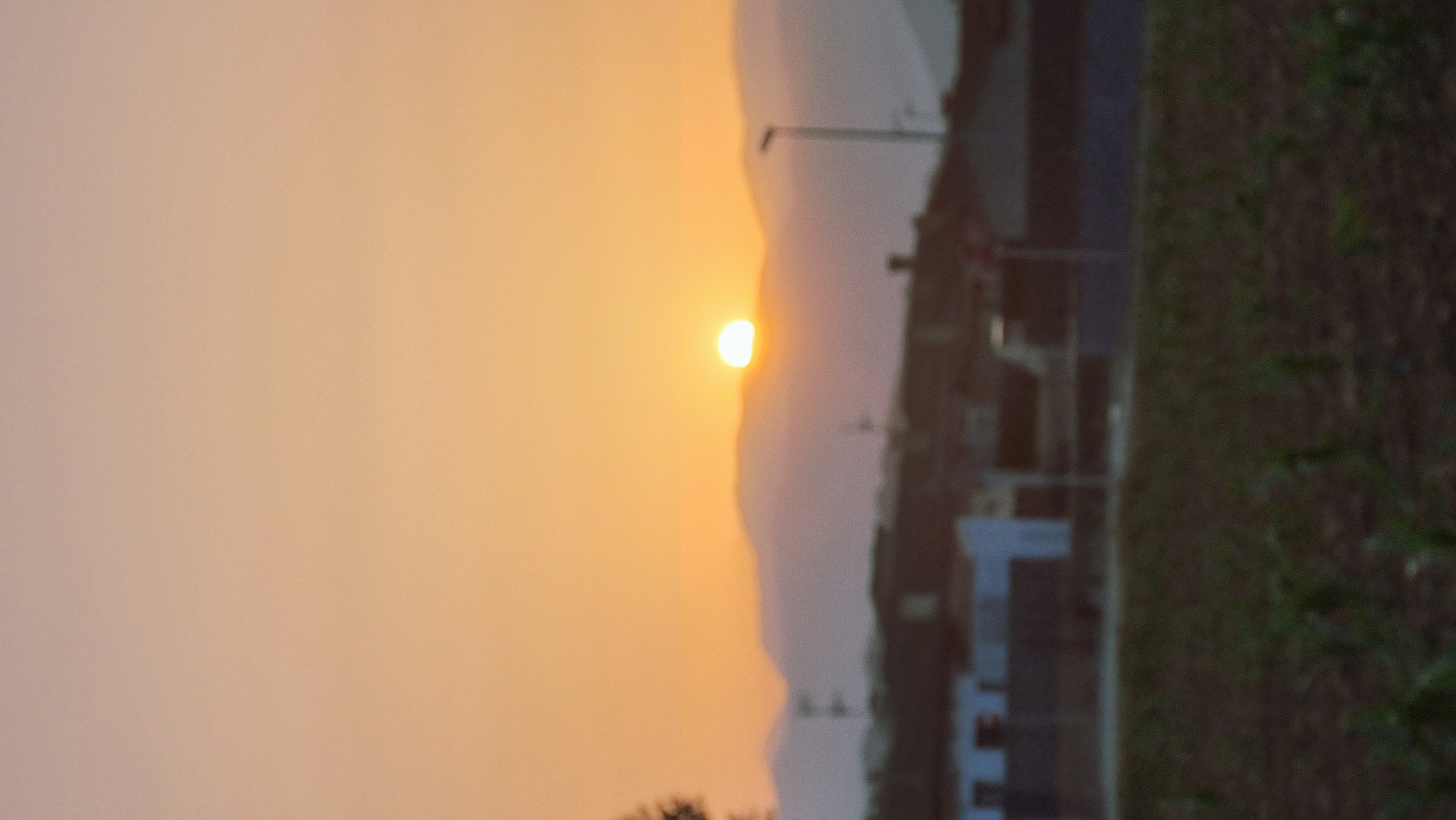 A landscape scene depicts a sunset over a mountainous horizon, with a partially visible sun emitting a warm glow. In the foreground, a blurry view of buildings and agricultural land is present. The scene is devoid of people or any discernible actions.