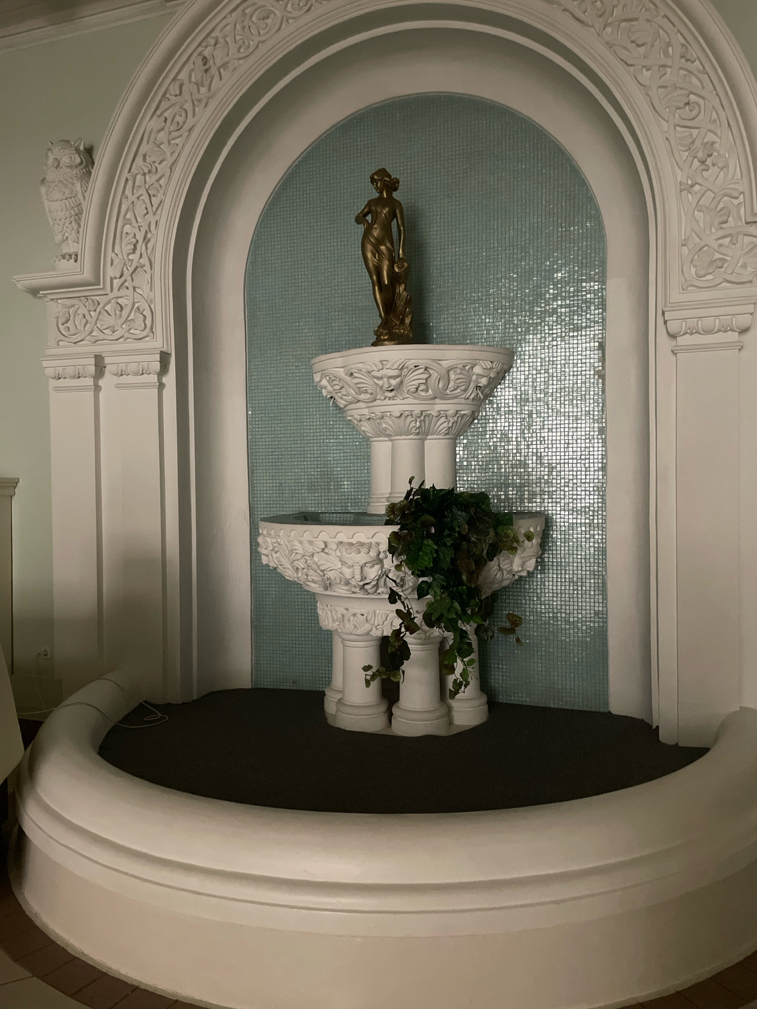 The image shows a decorative fountain situated within an arched alcove. The fountain is made of white plaster and features multiple tiers adorned with intricate carvings, including what appear to be faces and foliage. A golden statue of a female figure stands atop the upper tier. Green foliage spills over the side of the lower tier, adding a touch of nature to the artificial structure. The background of the alcove is covered in small, light blue tiles. The entire structure is framed by a larger, ornate white arch with decorative scrollwork and a small owl carving on the left side. The fountain sits on a dark carpeted surface, and in front of it is a curved, white plaster base. The lighting is dim, suggesting an indoor setting, possibly in a public space or a grand residence. The scene does not contain any people or any discernible indication of its location in Omsk, Russia.