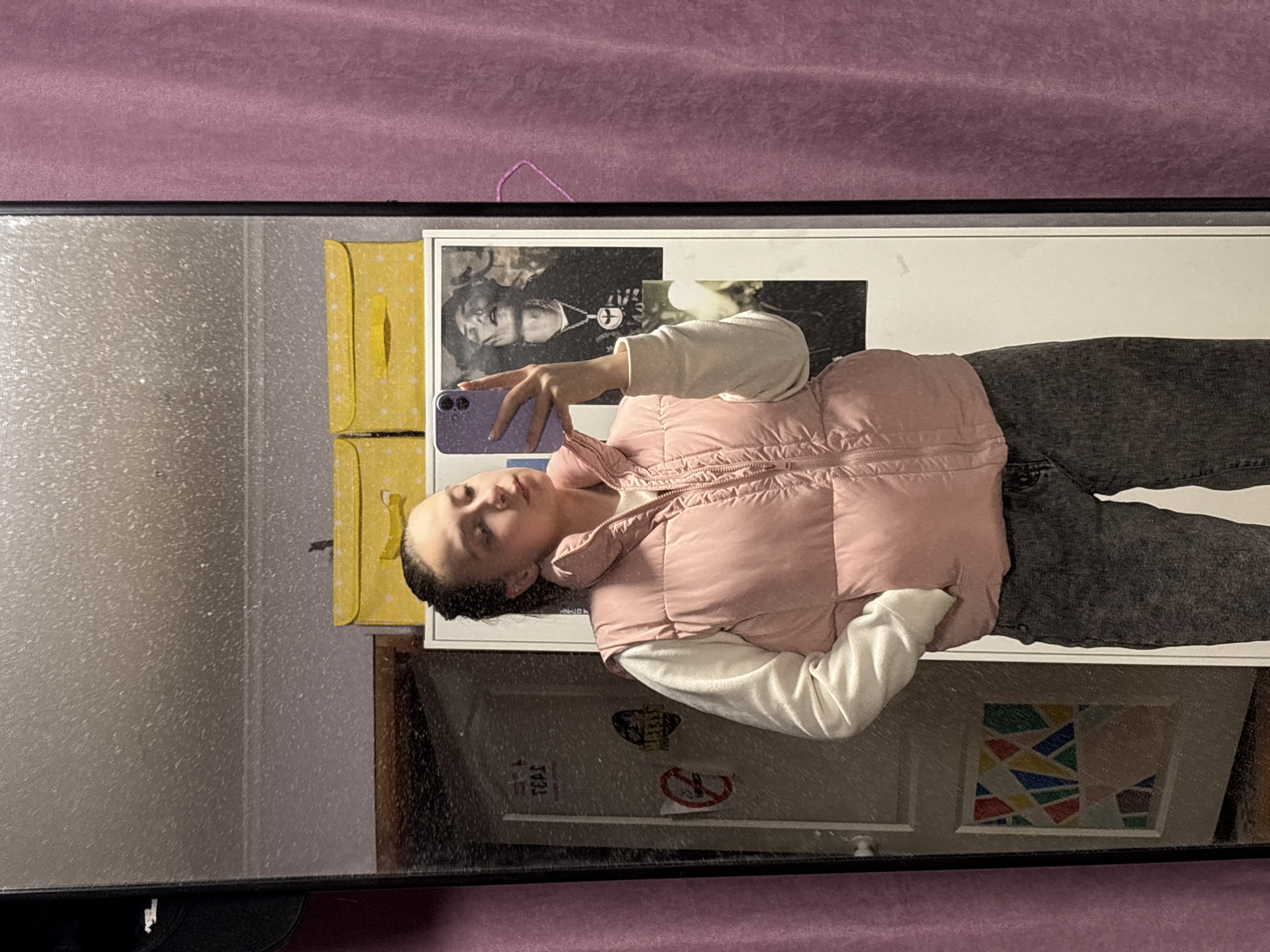 A mirror reflects a person taking a selfie. The person is wearing a pink vest, a white long-sleeved shirt, and gray pants. The reflection also shows a poster with a black and white image of a person, a yellow patterned box, and a door with a geometric art piece and stickers. The location is suggested to be Прикамский, Russia based on external information not visible in the image.