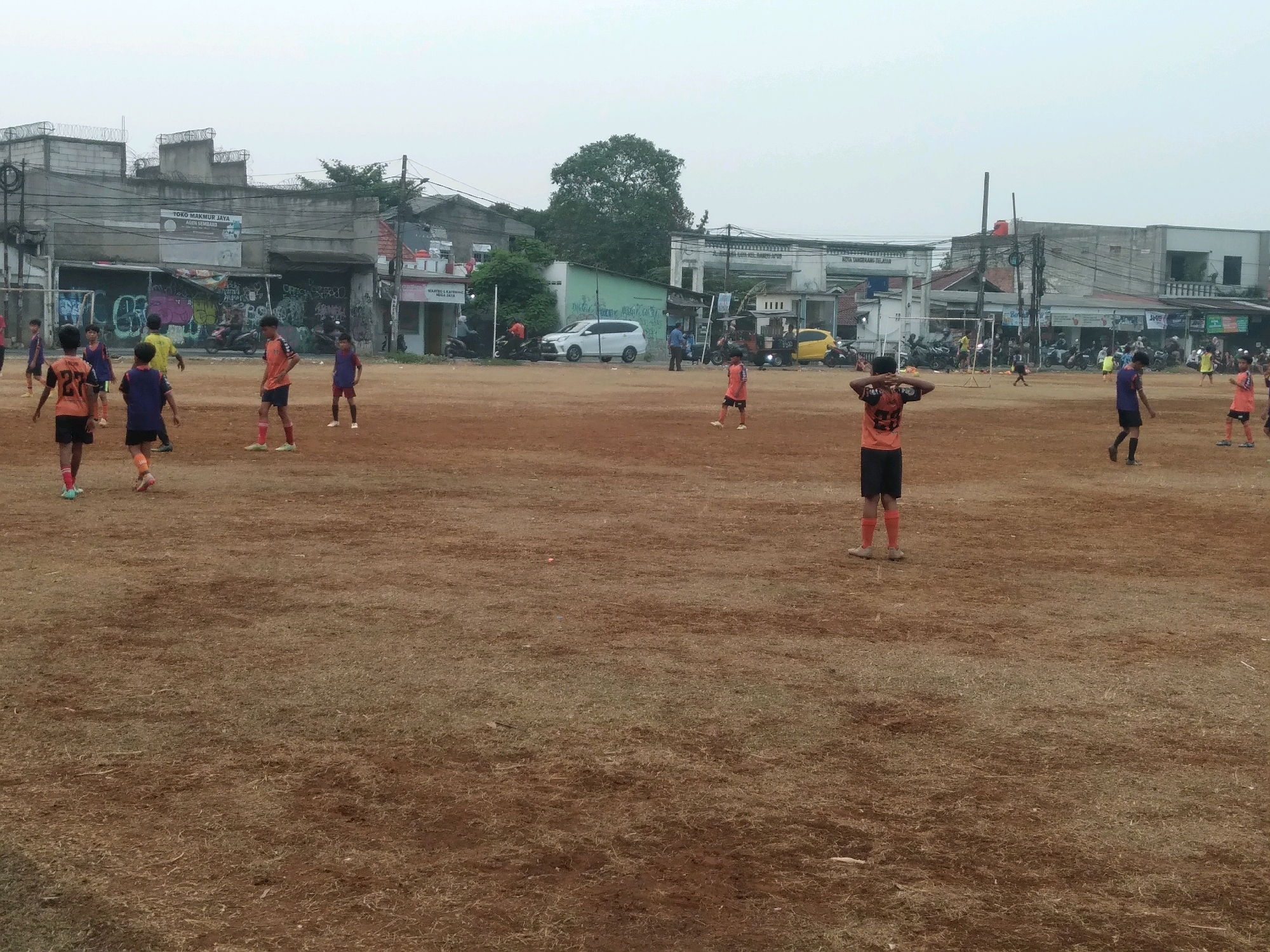 A group of young boys are playing soccer on a dirt field in an urban area of South Tangerang, Indonesia. The field is covered with dry grass and dirt, and there are buildings in the background. The sky is overcast. The boys are wearing soccer uniforms in various colors, including orange, blue, and purple. Some of the boys are running, while others are standing and watching. There is a soccer goal on the left side of the field. In the background, there are several buildings, some of which appear to be shops or houses. There are also some vehicles, including a white van and a yellow car. The overall scene depicts a casual soccer game taking place in a modest urban environment.