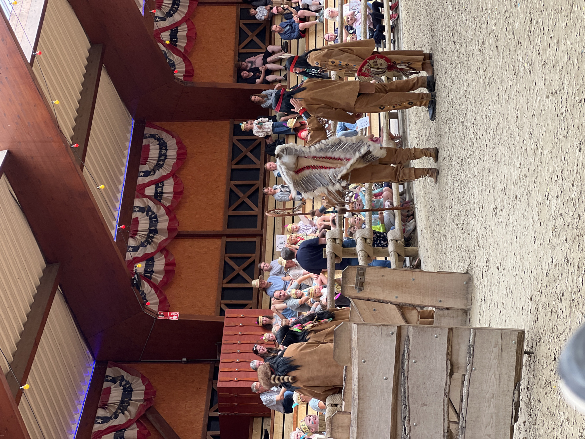 The image depicts an outdoor arena, likely within the city of Collendoorn, Netherlands, where a performance or reenactment is taking place. The scene is set in what appears to be a themed venue, possibly a Wild West attraction, judging by the attire of the main subjects.
In the foreground, three individuals are dressed in Native American-inspired costumes. The person on the left wears a brown buckskin outfit and a headdress adorned with fur and feathers. They are holding a large feathered shield-like object. The middle individual is also in brown buckskin, with a red headband and face paint. They appear to be gesturing towards the person on the right, who is wearing a similar buckskin outfit with intricate beadwork and a feathered ornament. The ground of the arena is covered in sand or fine gravel.
Behind the performers, wooden barricades and fencing are visible, separating the performance area from the audience. A crowd of spectators is seated on tiered wooden benches in the background. They are of various ages and are casually dressed. Some are looking towards the performance, while others are engaged in conversation.
The backdrop of the arena is a wooden structure with large, dark-stained beams and plywood walls. American-style bunting, featuring red, white, and blue stripes with stars, is draped along the upper edges of the structure, suggesting a patriotic or thematic element to the venue. Strings of colored lights, some red and some yellow, are strung across the ceiling beams. The overall atmosphere appears to be that of a family-friendly entertainment event.