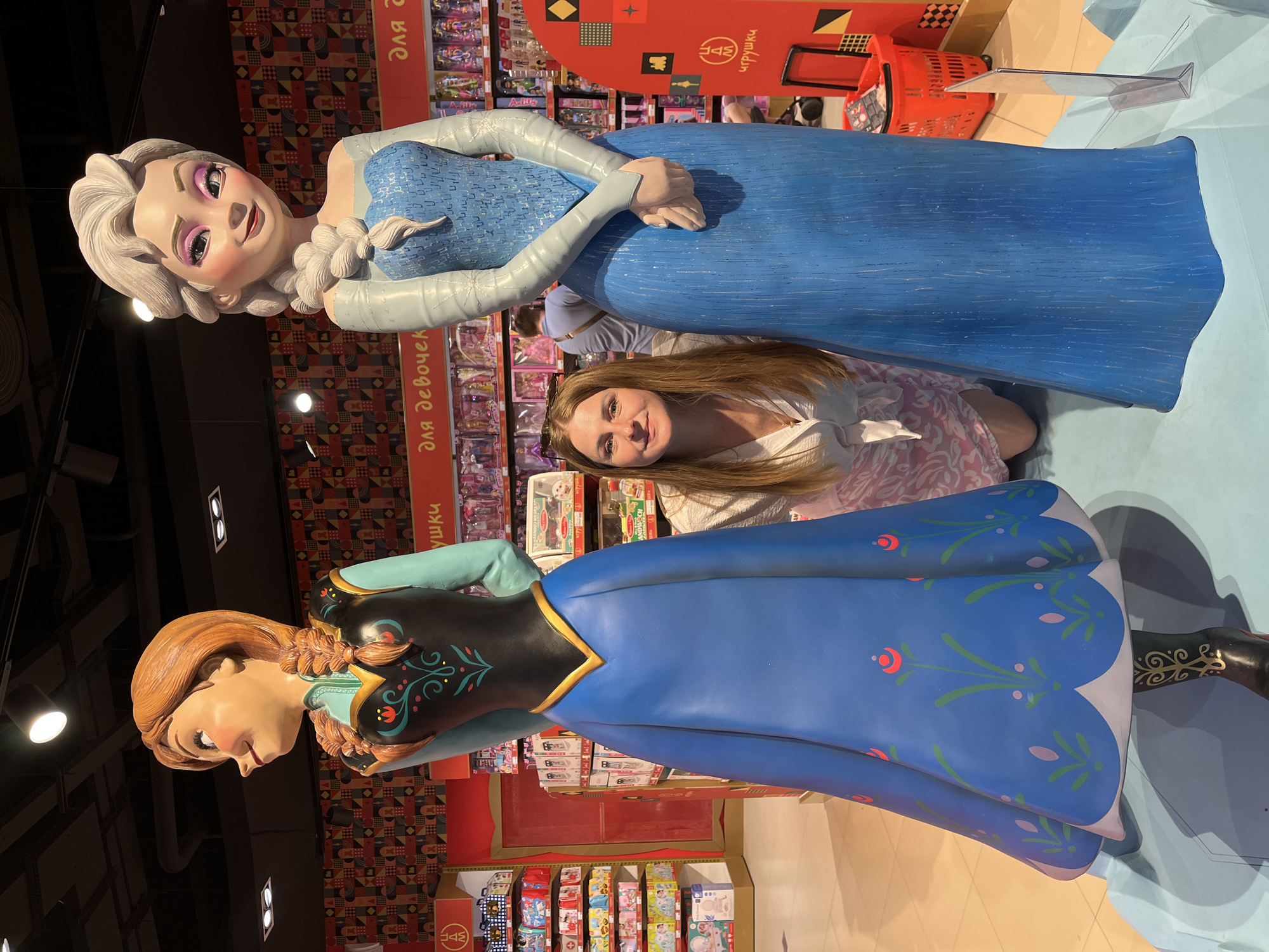 An adult female kneels between two life-sized statues of Disney's Princess Anna and Queen Elsa within an indoor commercial space, likely a toy store, in a Russian-speaking country (city of Unknown, Unknown).
The woman, with long light brown hair and wearing a light-colored collared shirt and patterned light pink and white skirt, is positioned centrally, smiling directly at the viewer. She has sunglasses resting on her head.
To her left stands the Anna statue, depicted with reddish-brown braided pigtails. She wears a light blue long-sleeved shirt beneath a black bodice adorned with red, green, and pink floral embroidery, and a floor-length royal blue skirt featuring similar floral patterns and a white scalloped hem, with visible black boots accented with gold. Anna's head is turned slightly to her left with a gentle smile.
To her right stands the Elsa statue, featuring platinum blonde hair in a long braid over her left shoulder. She is dressed in a sparkling ice-blue gown with an off-the-shoulder top and a textured bodice, transitioning into a plain blue skirt. Her hands are clasped in front of her, and she is turned slightly to her right with a soft smile.
Both statues stand on a light blue platform. The background consists of tall shelves stocked with various children's toys and merchandise. Red banners with white Russian text reading "ДЛЯ ДЕВОЧЕК" (FOR GIRLS) and "ИГРУШКИ" (TOYS) are prominently displayed. A partial view of an adult male in a light blue shirt is visible behind the Elsa statue. The environment features patterned wall panels, a dark ceiling with spotlights, and a light-colored floor. A red shopping basket and a red display stand with Russian text are present on the right side of the frame.
