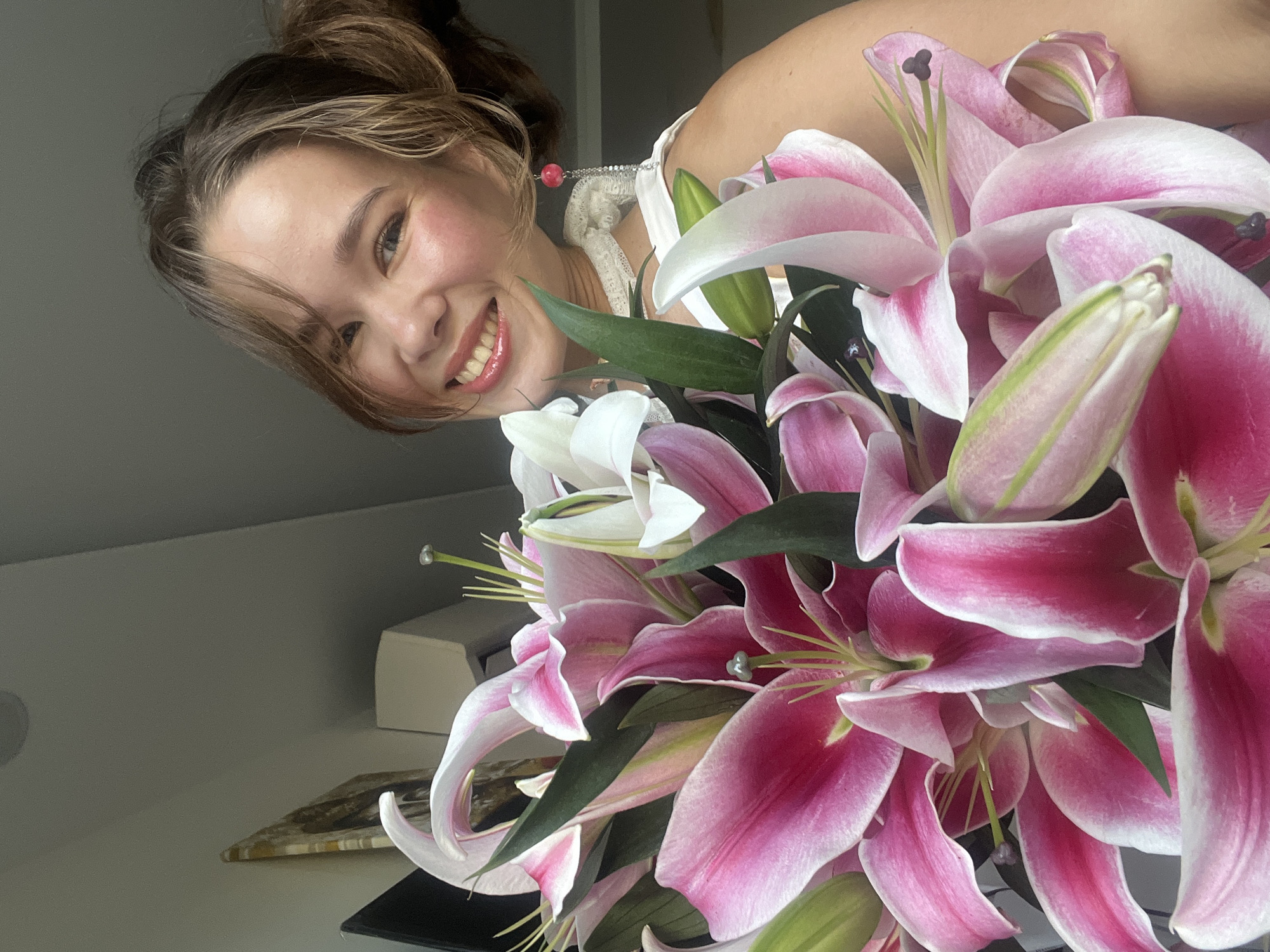 A woman smiles while holding a bouquet of pink and white lilies. She is wearing a white top and an earring with a red bead. The background shows a corner of a room with a white shelf holding various objects. The location of Nam Natural Heritage Area Trang, Vietnam is not discernible from the provided image.