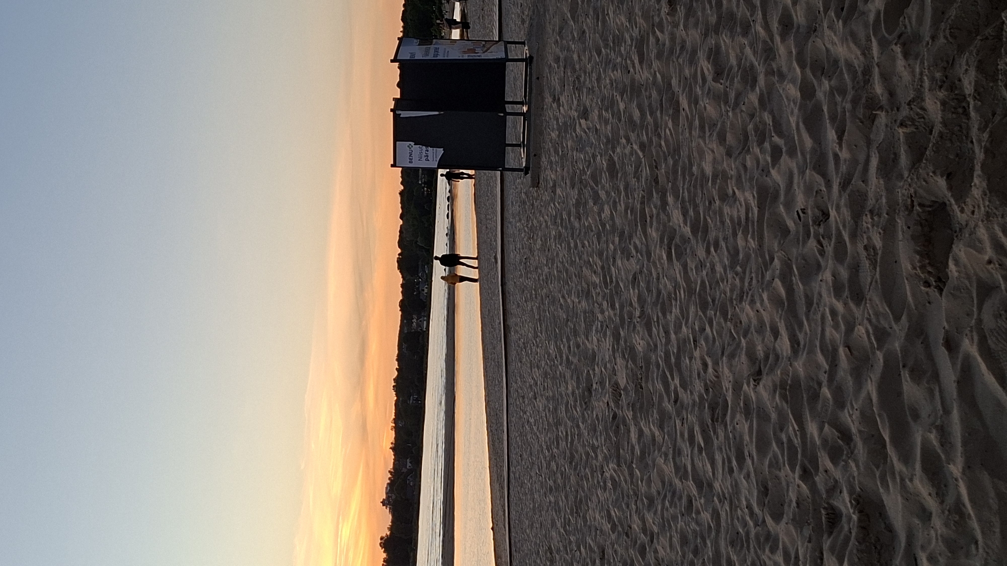 The image depicts a beach scene at sunset in Tallinn, Estonia, likely at Pirita Beach. In the foreground, the sandy beach is textured with ripples. In the middle ground, two individuals are walking near the water's edge, silhouetted against the warm, orange glow of the sunset sky. Further to the right, a black structure, possibly a changing room or information booth, stands on the sand. The background shows a body of water reflecting the sky, with a treeline and buildings visible on the distant shore. The overall atmosphere is serene, with soft lighting characteristic of twilight.