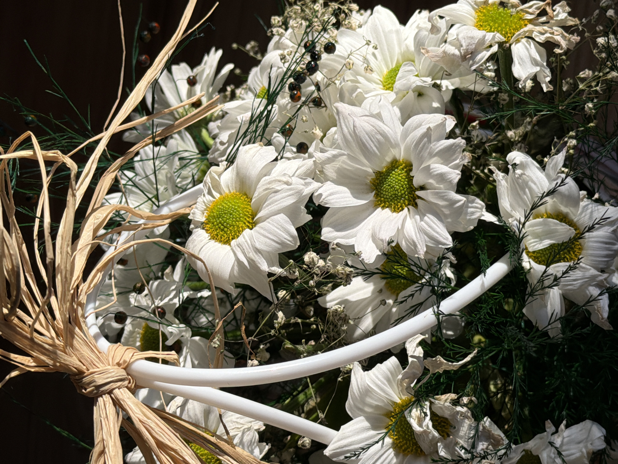 A close-up shows a floral arrangement with white daisy-like flowers featuring green centers. The arrangement is partially obscured by a white, tube-like structure, and tied with a bundle of raffia. Dried plant matter, including fine stems and seed heads, is interspersed among the flowers. The background is dark, creating high contrast with the light-colored flowers and elements of the arrangement. No people or specific identifiable location in the city of Unknown, Unknown are apparent in this image.