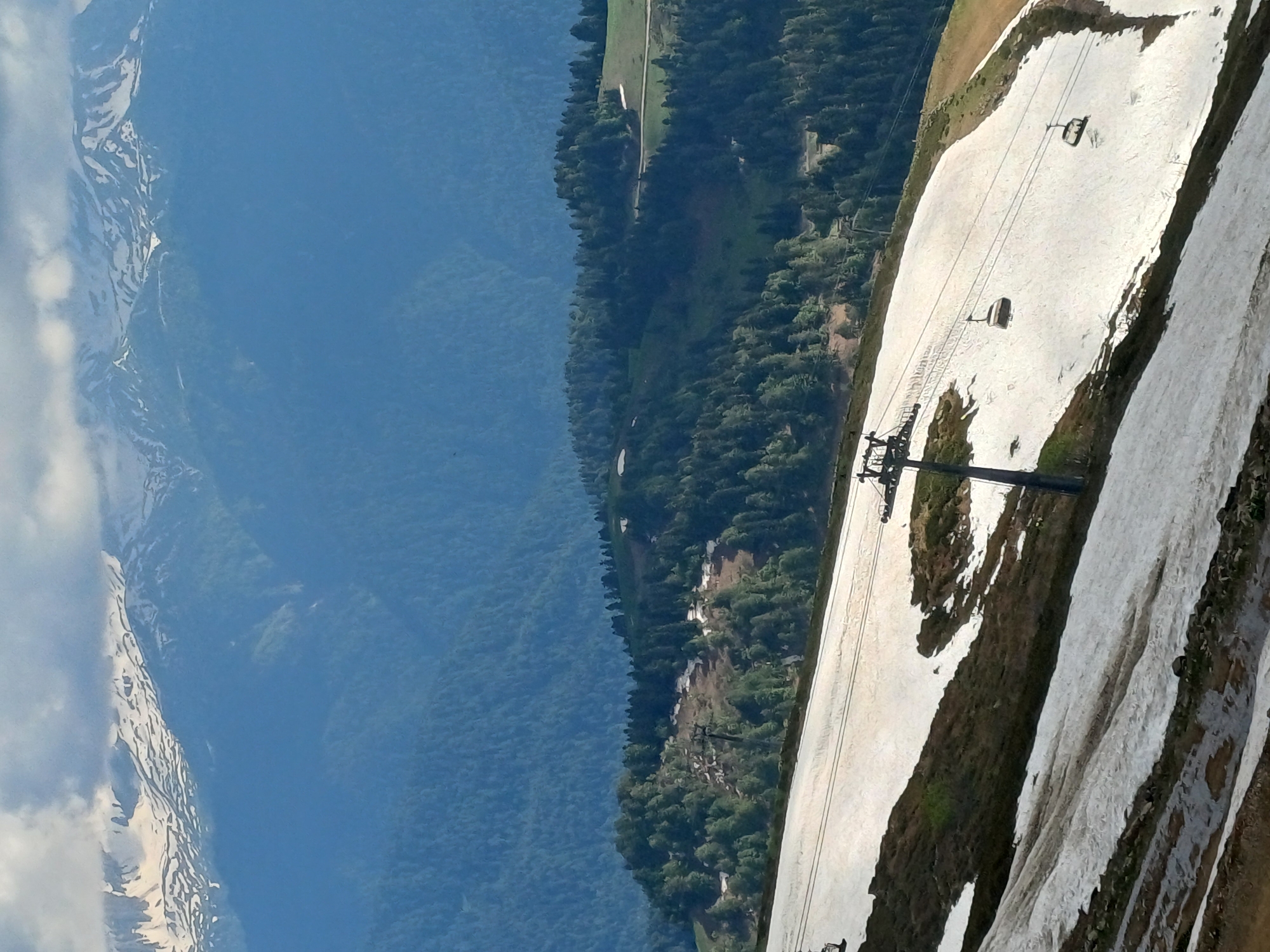 This is a tilted, medium-shot photograph taken from a high vantage point, looking down at a ski resort in Estosadok, Russia. The scene is dominated by a ski slope, partially covered in snow, that stretches diagonally across the frame from the upper right to the lower left. A ski lift with chairs suspended on a cable runs parallel to the slope, with two chairs visible. The surrounding landscape features lush green mountains and dense forests. The sky is a clear, bright blue, suggesting it is daytime with good weather. Patches of snow are scattered across the mountainside, indicating a transition between seasons. The overall impression is one of natural beauty and recreational activity. There is no visible text in the image.