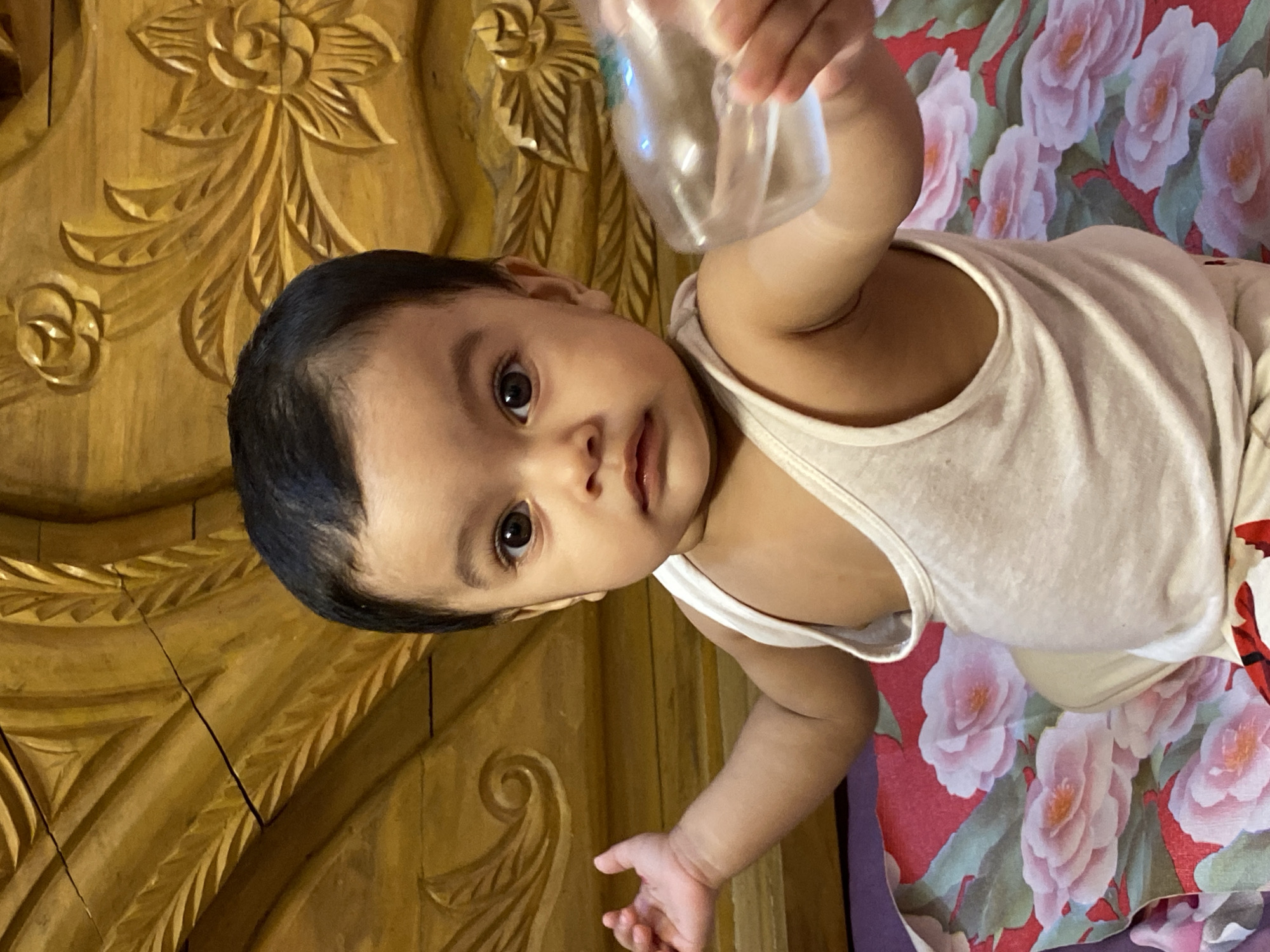 An overhead shot captures a baby looking directly at the camera, holding a clear plastic bottle in their raised hand. The baby appears to be an infant or young toddler, dressed in a simple white sleeveless top or vest, and has dark hair and brown eyes. The plastic bottle is semi-transparent, reflecting some light.
The baby is situated on a surface that is part wood and part patterned fabric. To the baby's left, a section of rich, golden-brown wooden furniture, likely a headboard or bed frame, is visible. It features intricate carvings of floral designs, including large roses and leaves, indicating traditional craftsmanship. To the baby's right and beneath them, there's a vibrant red fabric with a pattern of large, soft pink roses and green leaves, possibly a bedsheet or blanket.
The scene takes place indoors, likely in a bedroom, during the daytime, suggested by the natural lighting. The baby's expression is calm and inquisitive, directly engaging with the viewer. There is no visible text in the image. The style of the carved wooden furniture and bedding is consistent with a domestic setting in Dhaka, Bangladesh.